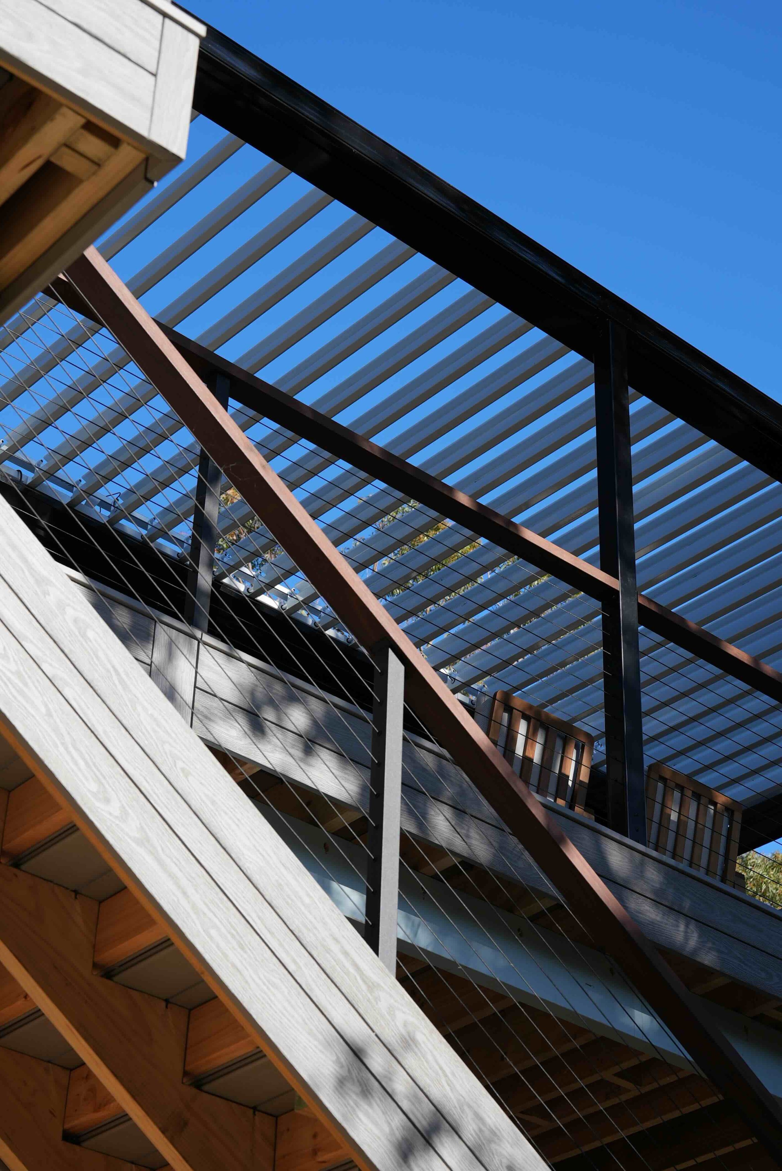 View of a modern outdoor deck with wooden and metal railings, under a bright blue sky with some trees visible in the background.