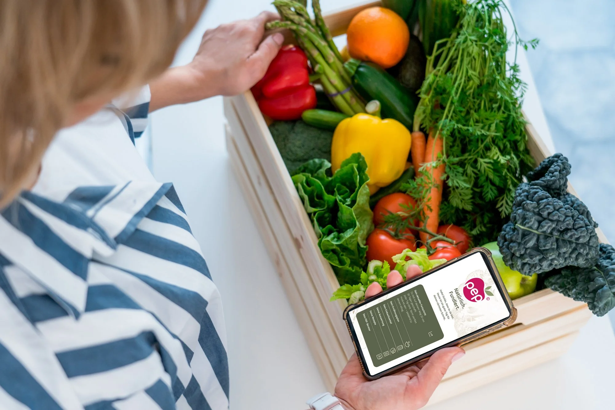 Frau hält Smartphone mit der pep-Ernährungsprogramm App über pflanzenbasierte Ernährung vor einem Korb mit frischem Gemüse, darunter Paprika, Gurke, Karotten, Rote Beete und grünes Gemüse, an einer weißen Oberfläche.