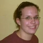 Eine junge Frau mit braunen, lockigen Haaren, trägt eine Brille und lächelt vor einem hellgelben Hintergrund.