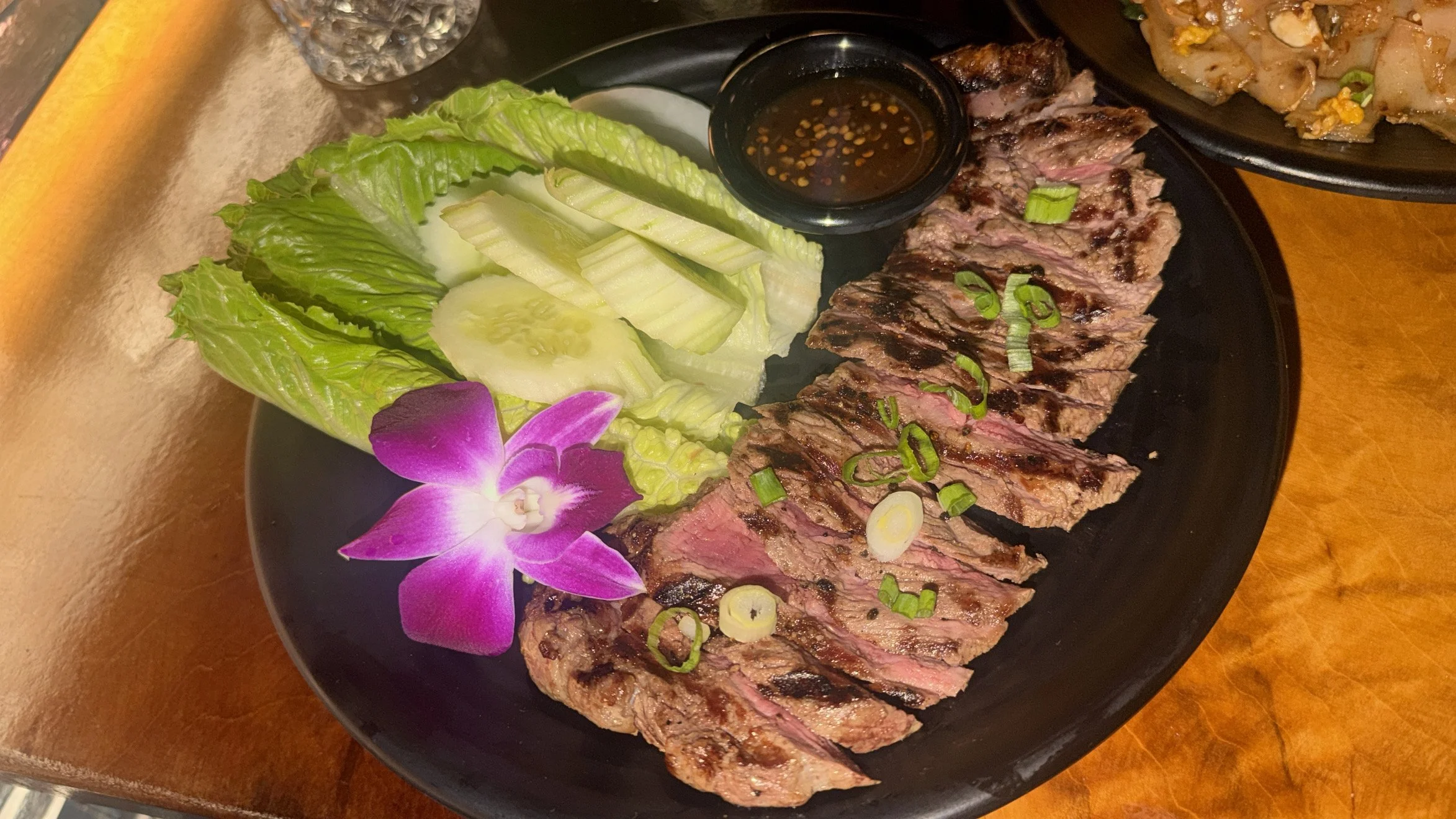 Sliced grilled beef steak with green onions, arranged on a black plate with lettuce, cucumber slices, and a purple orchid flower, accompanied by a small bowl of dipping sauce, on a wooden table.