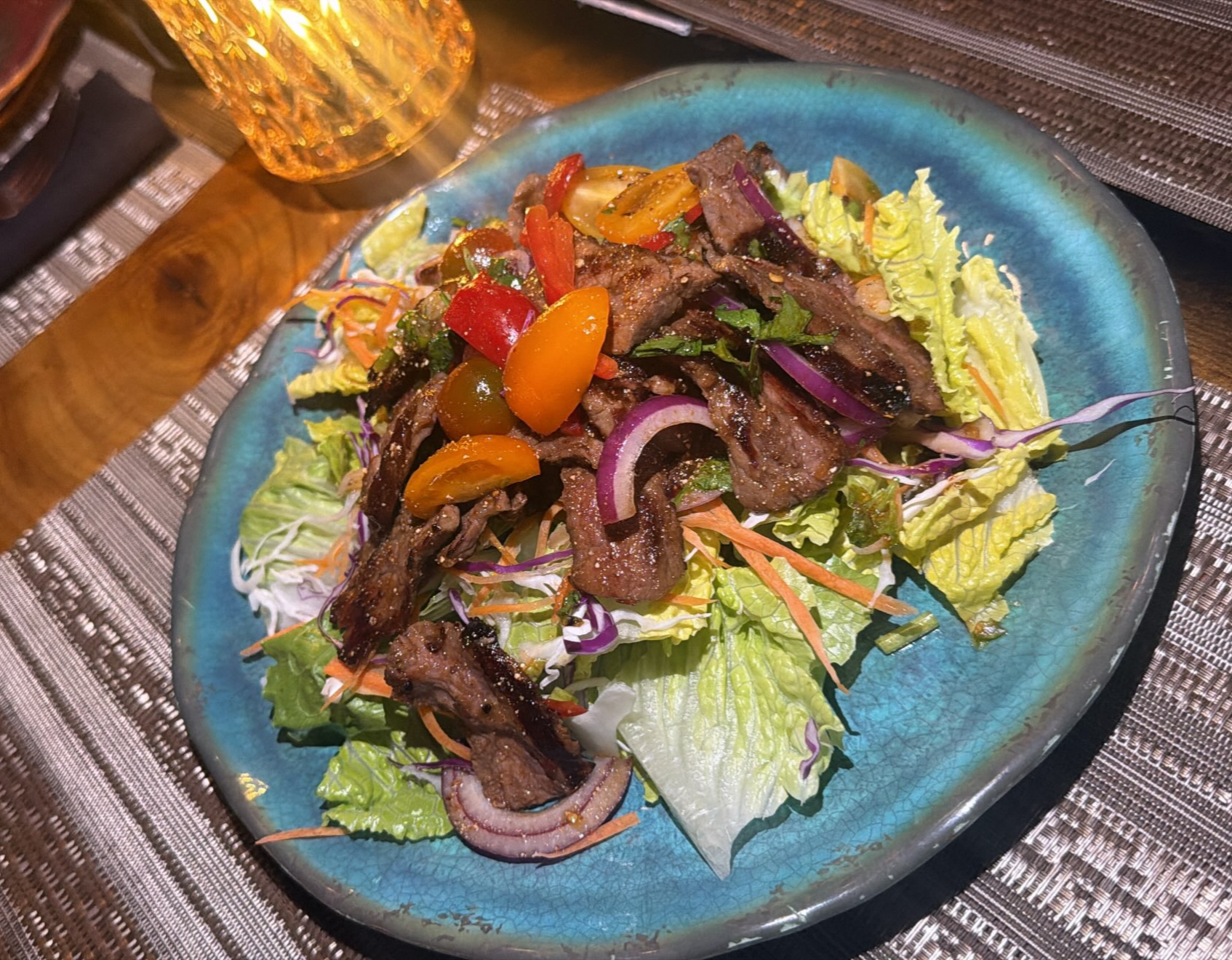 A plate of grilled steak strips on a bed of mixed salad greens, shredded carrots, sliced red onions, cherry tomatoes, and chopped cilantro on a blue ceramic dish with a glass of amber-colored beverage in the background.