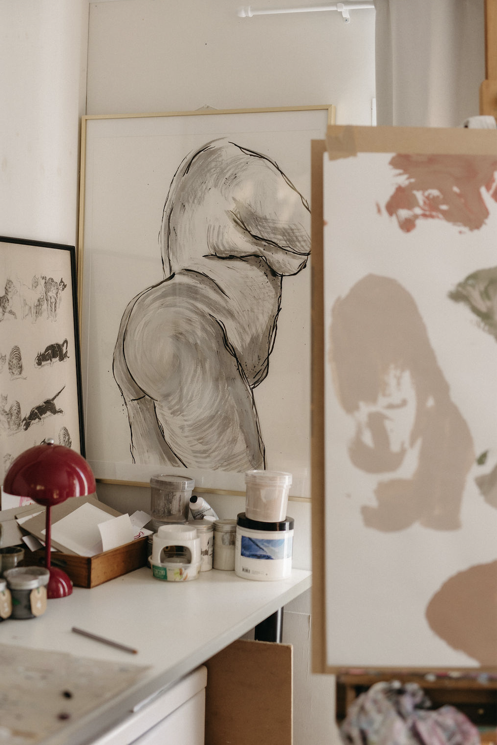 Artist's studio with framed figure drawings, paint jars, brushes, and red desk lamps.