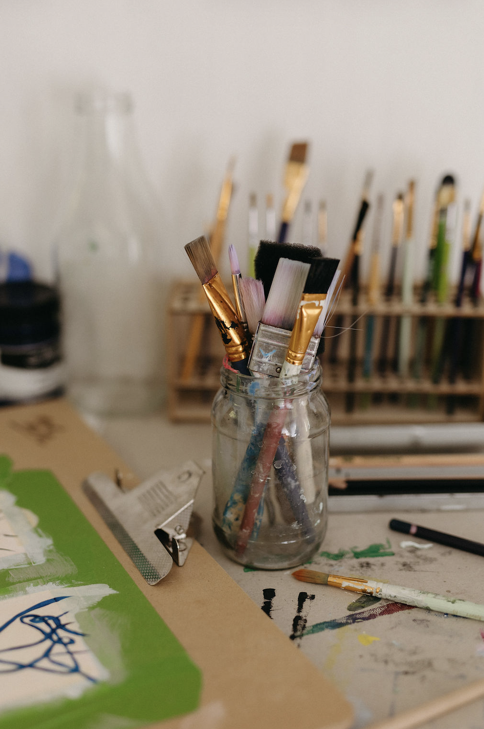 Jar of assorted paintbrushes on an artist’s worktable beside paint-smeared artwork and supplies