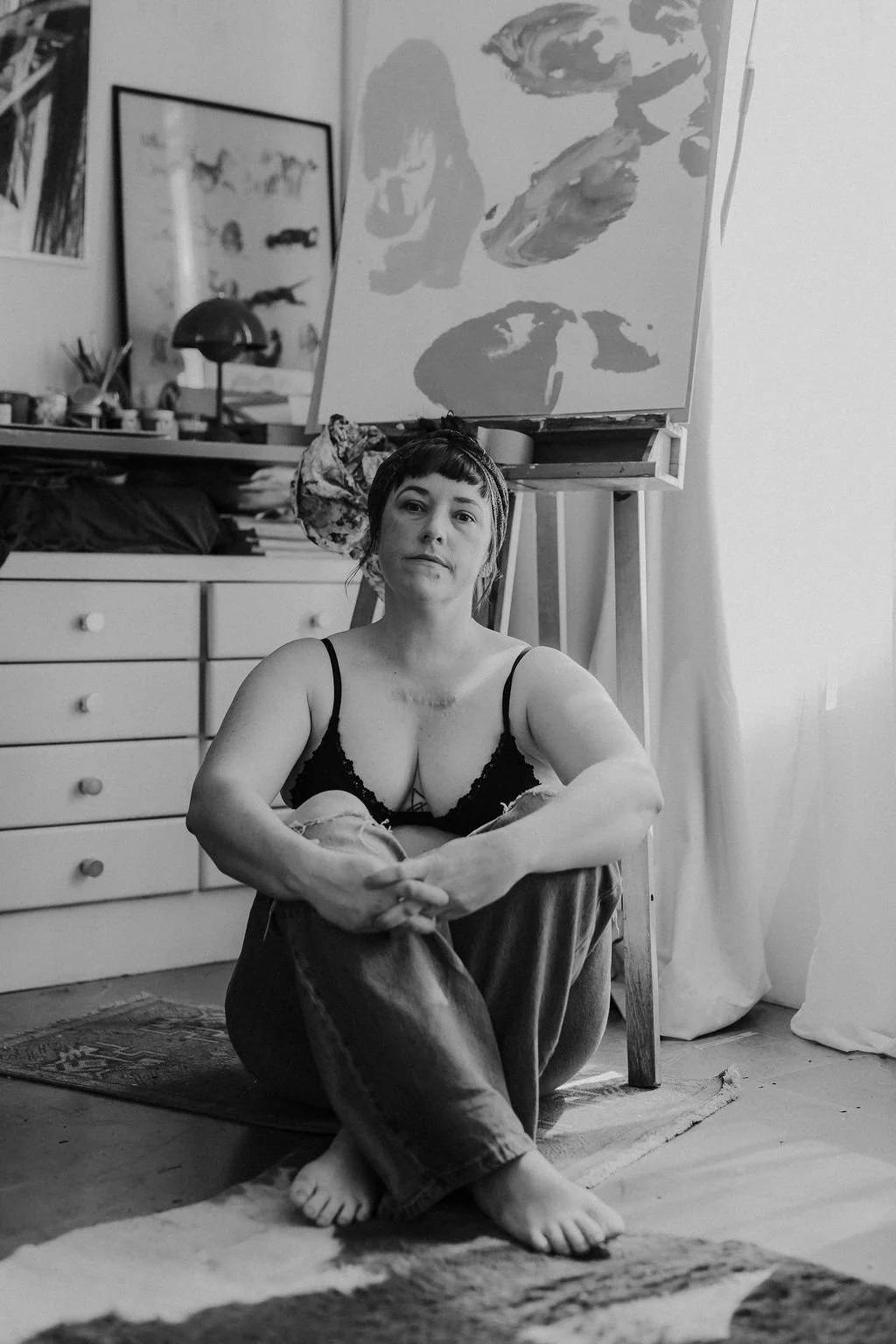 Black-and-white portrait of a woman sitting barefoot on the floor in an art studio, with paintings and furniture behind her.