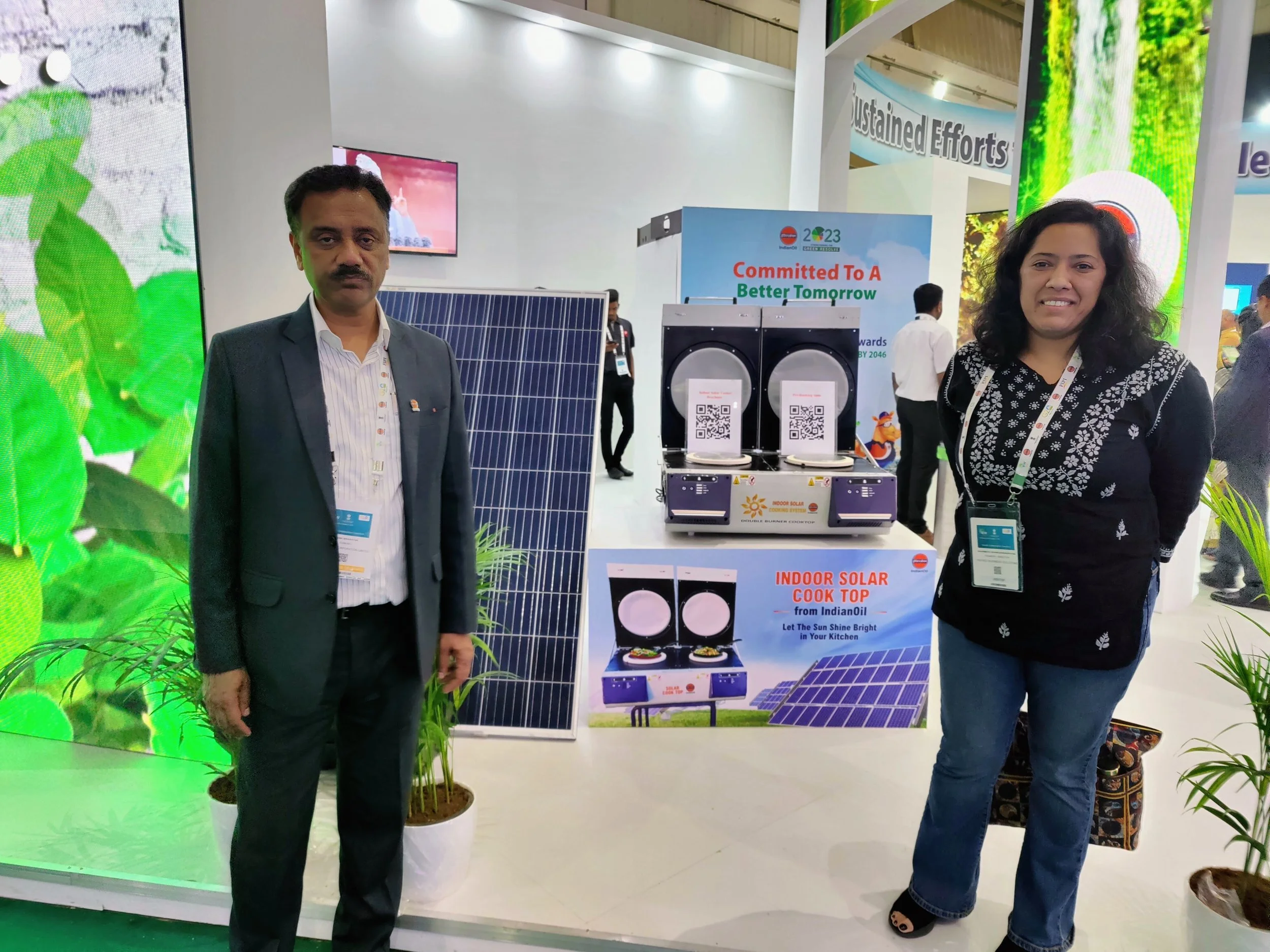Two people standing in front of a booth at a trade show. The man on the left is wearing a dark blazer and light shirt, and the woman on the right is wearing a black patterned top and jeans. Behind them, there are solar panels, a sign that reads "Committed To A Better Tomorrow," and a display promoting indoor solar cooking from IndianaOil. Green plants are positioned around the booth.