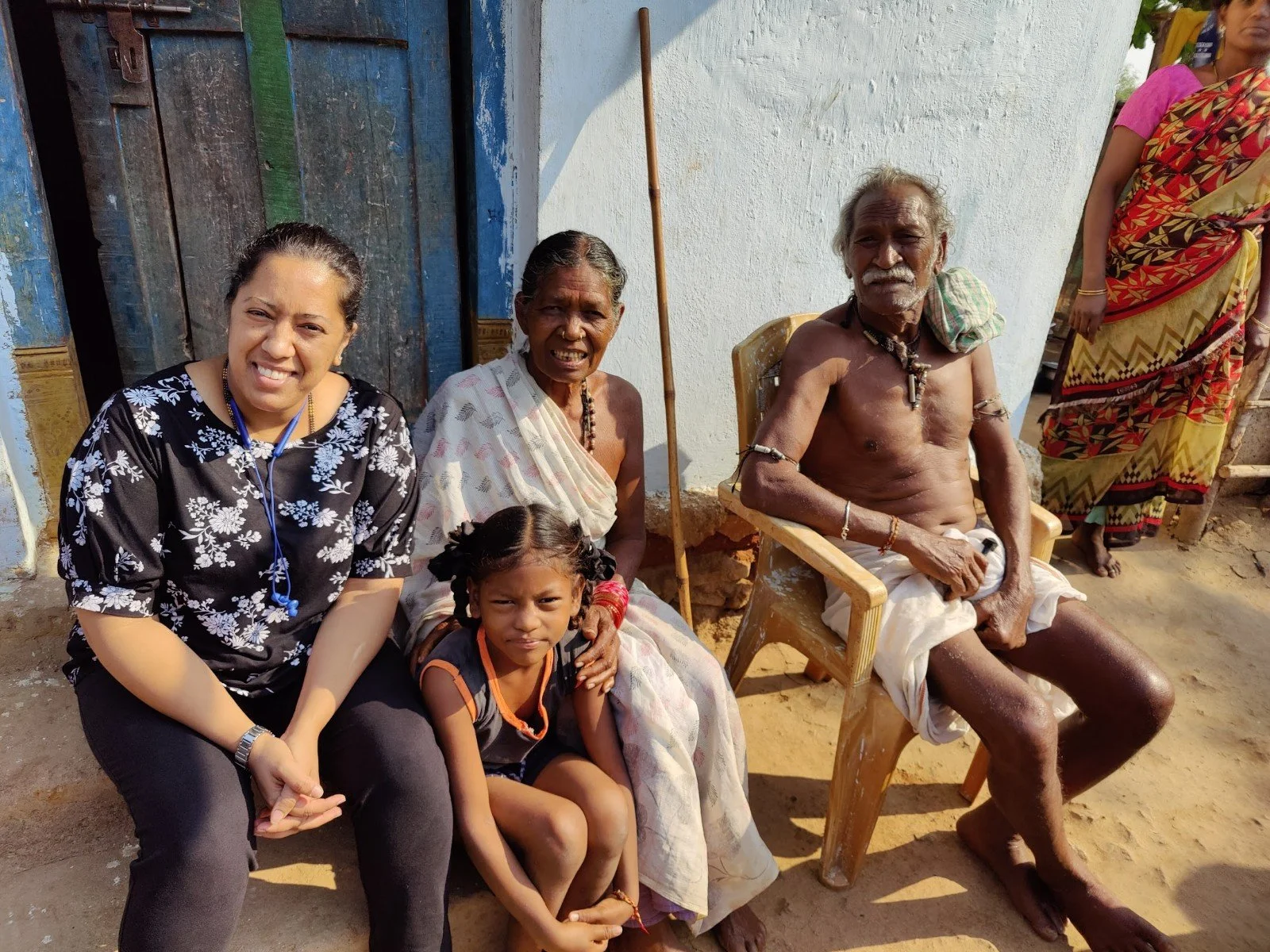 A group of five people, including a young girl, two older women, a shirtless elderly man, and a woman standing, sitting outside a building with a blue door. Some are seated on chairs, and the background shows a white wall and outdoor environment.