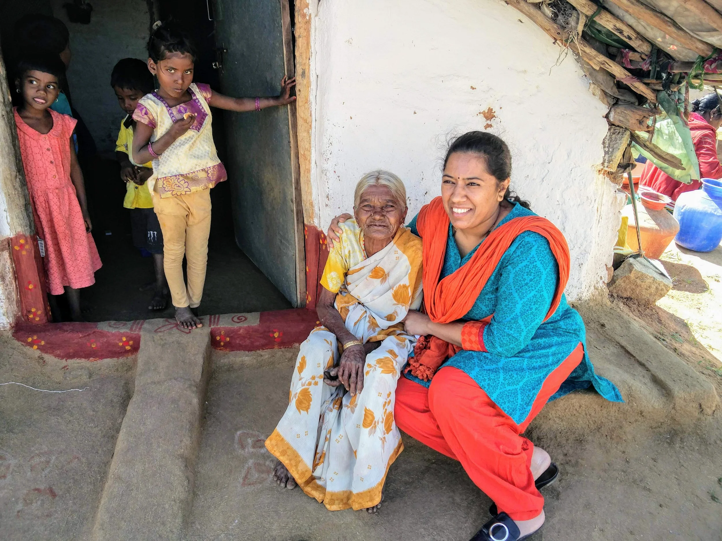 A group of children and two women outside a simple house with a white wall and thatched roof, sitting or standing on a concrete step; they are casually dressed, with some children looking at the camera and the women smiling.
