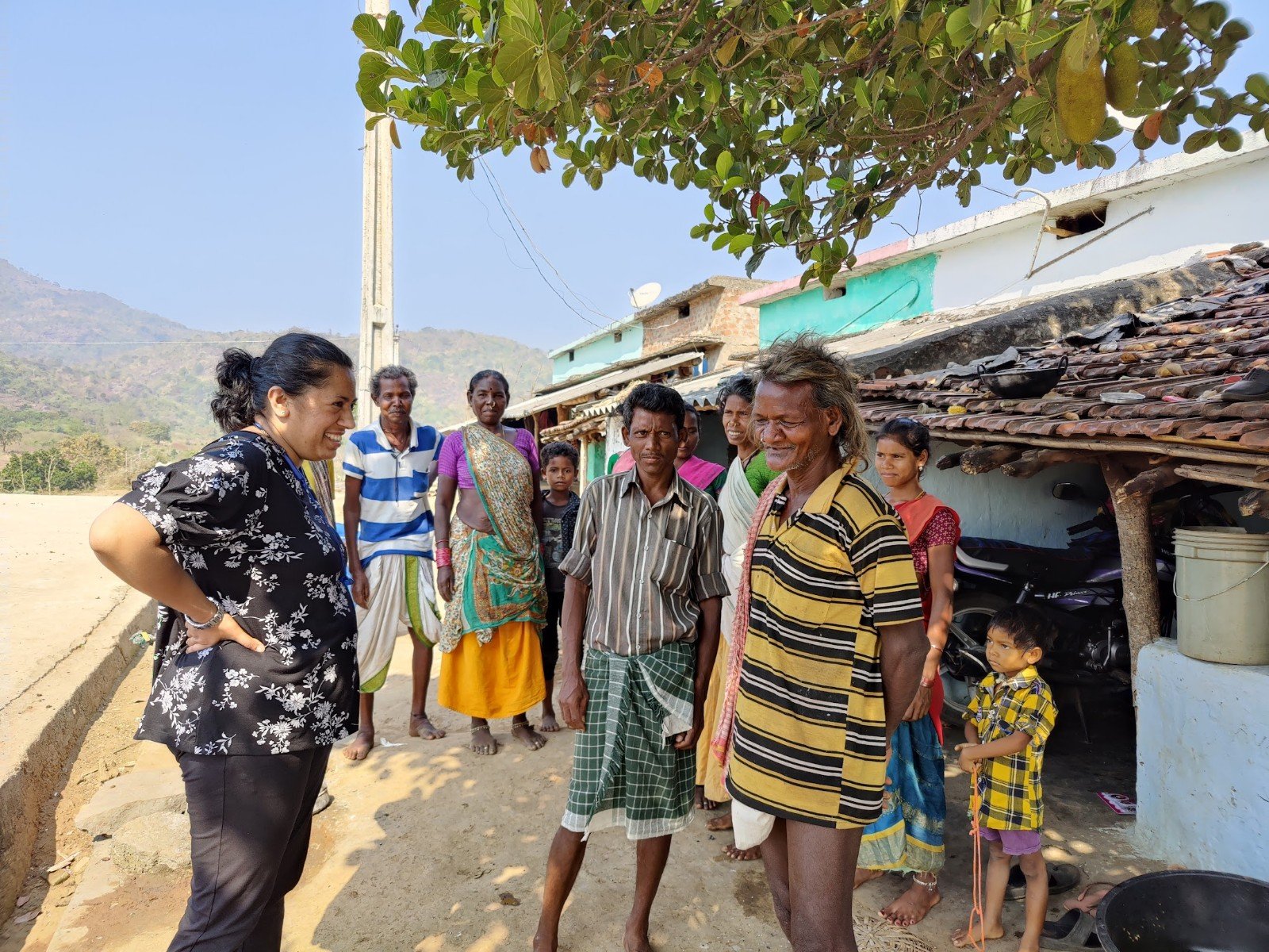 A group of people, including men, women, and children, gathered outdoors in a village setting, engaged in conversation with a woman in a black floral shirt. There are houses with tiled roofs, a tree, and mountains in the background.