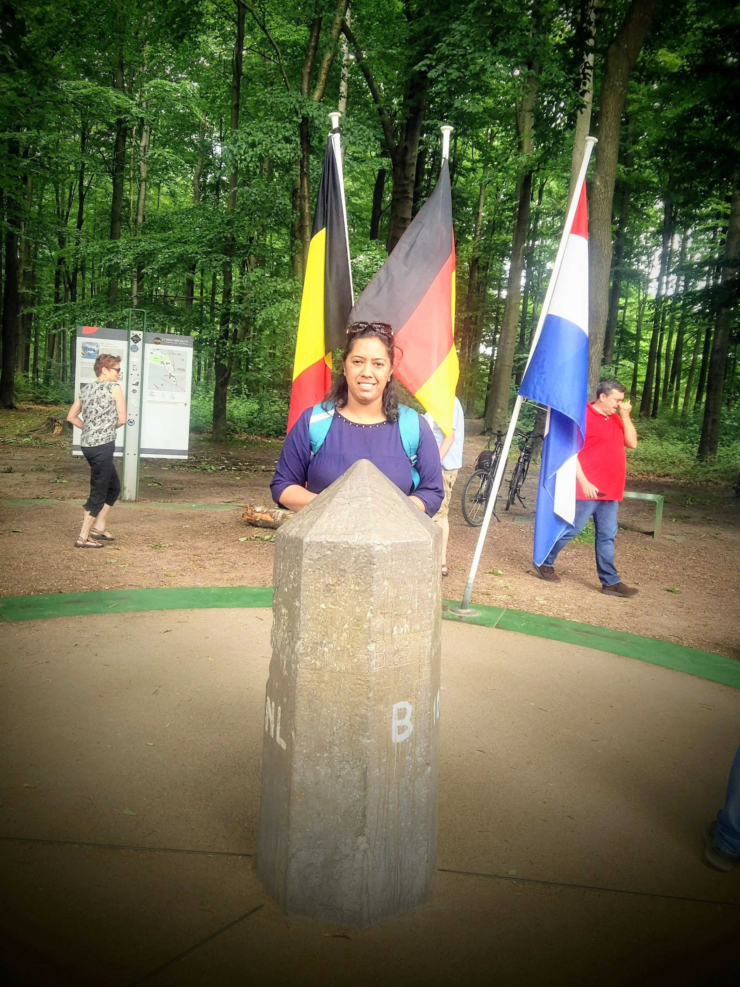 A woman with a backpack standing behind a stone boundary with the letter 'B' engraved on it. Behind her are three flags, including the German, French, and Dutch flags, in a wooded park with trees and a trail map.