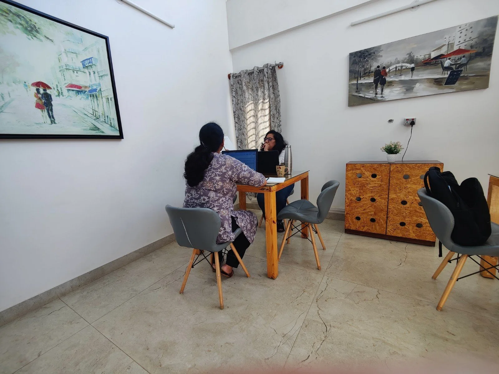 Two women sitting at a small wooden table in an office or meeting room, engaged in conversation, with laptops and coffee mugs. Artwork hangs on the white walls, and there is a wooden cabinet with a potted plant on top.