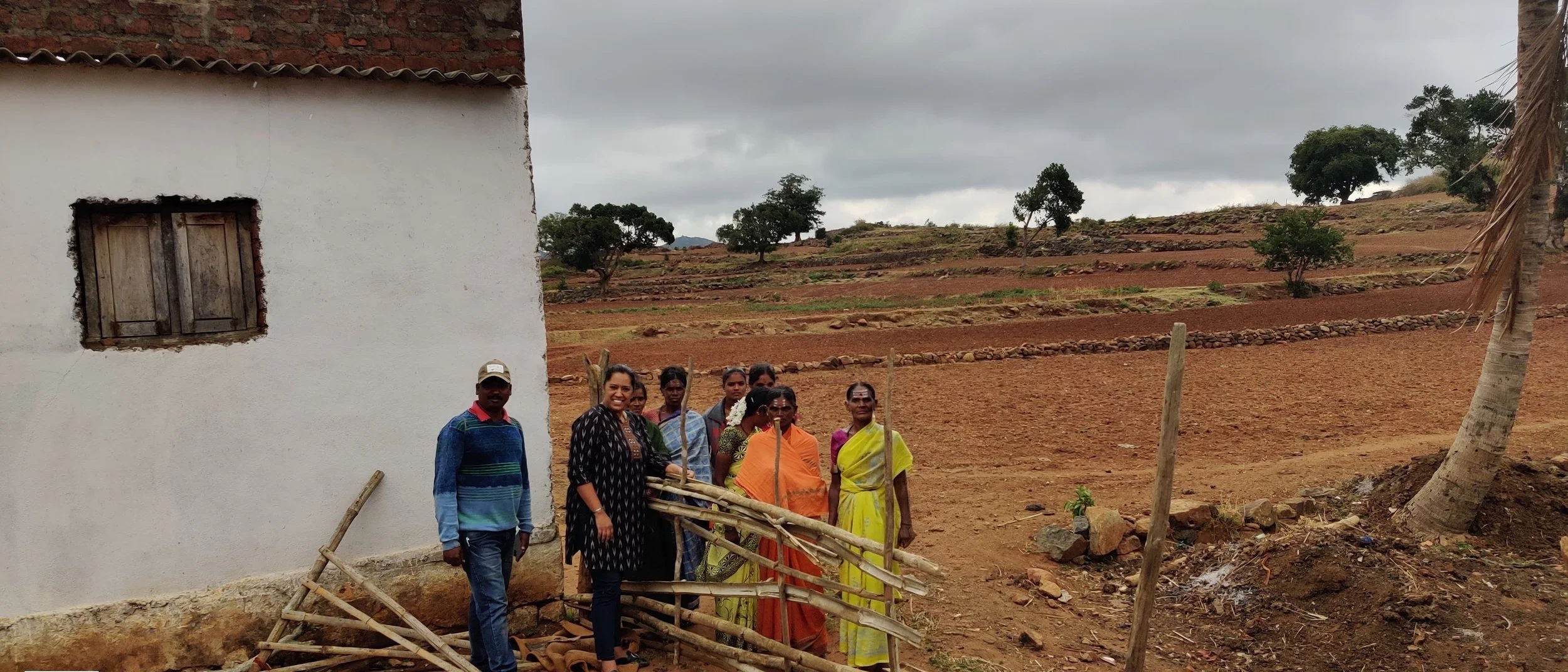 A group of people standing outside near a white building with a small window. The landscape features terraced red soil fields and scattered trees under a cloudy sky.
