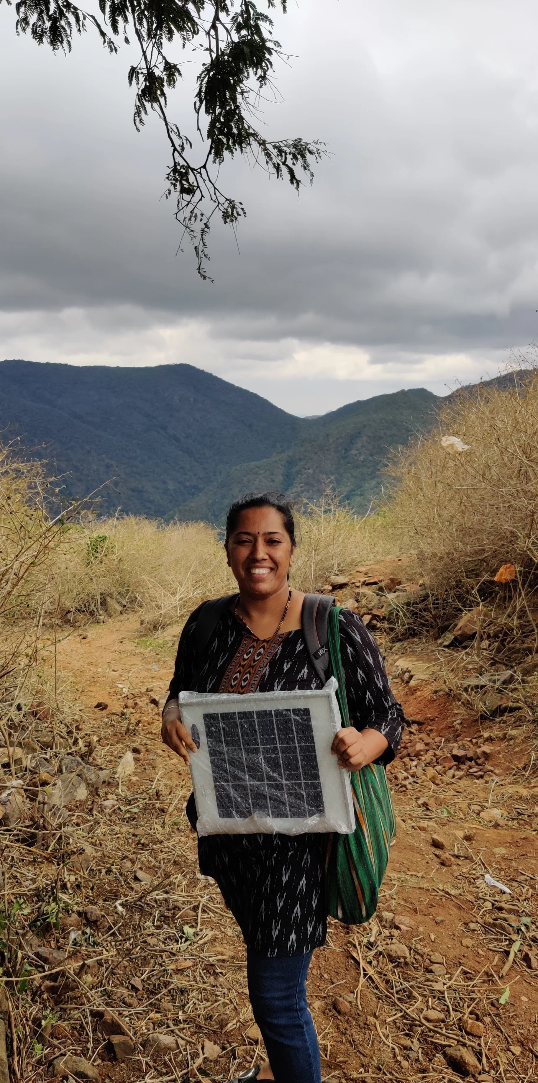 A woman smiling in a natural outdoor setting with mountains and cloudy sky in the background, holding a solar panel.