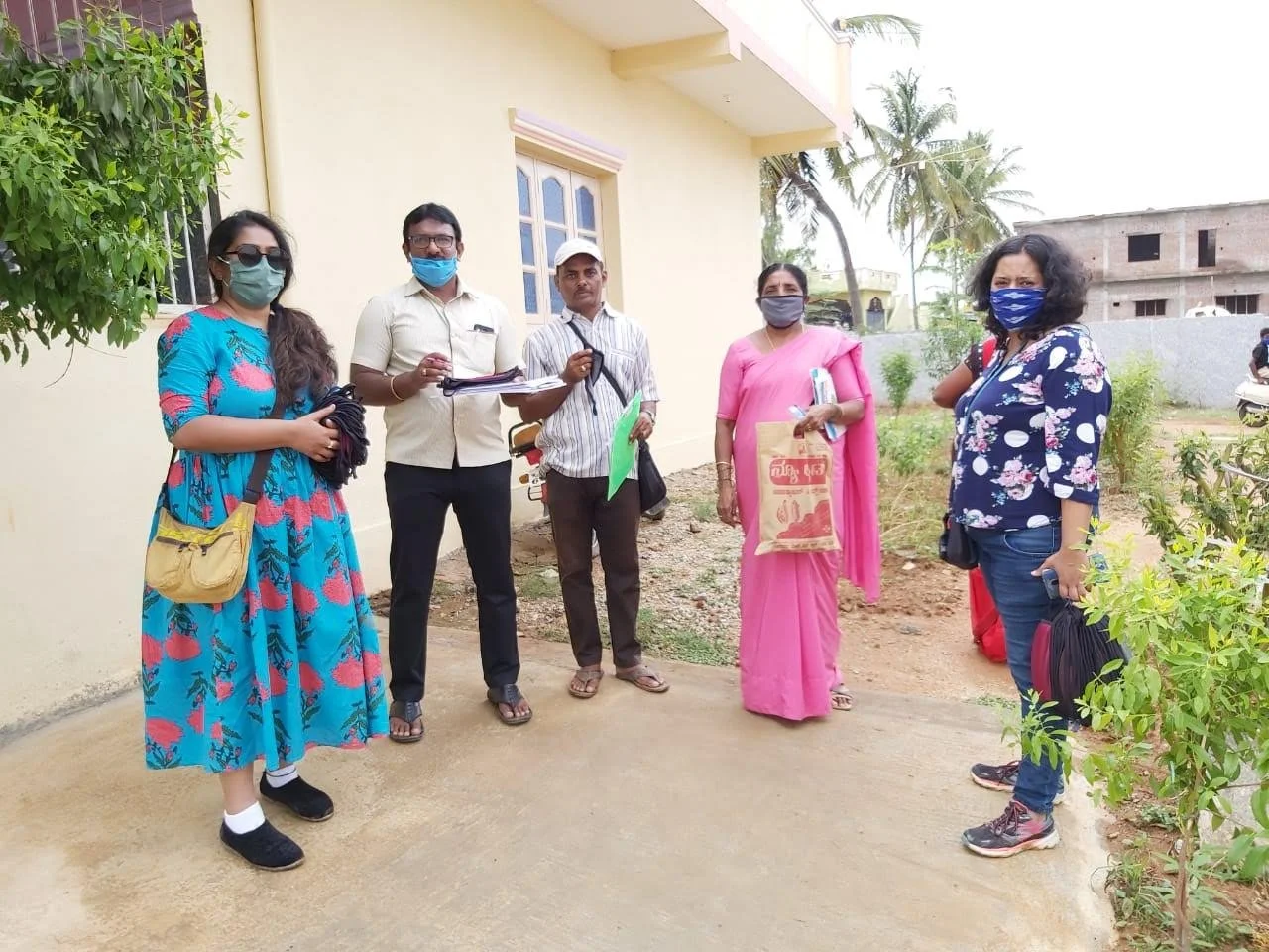 Five people standing outdoors near a light-colored building, some holding bags and documents, all wearing masks.