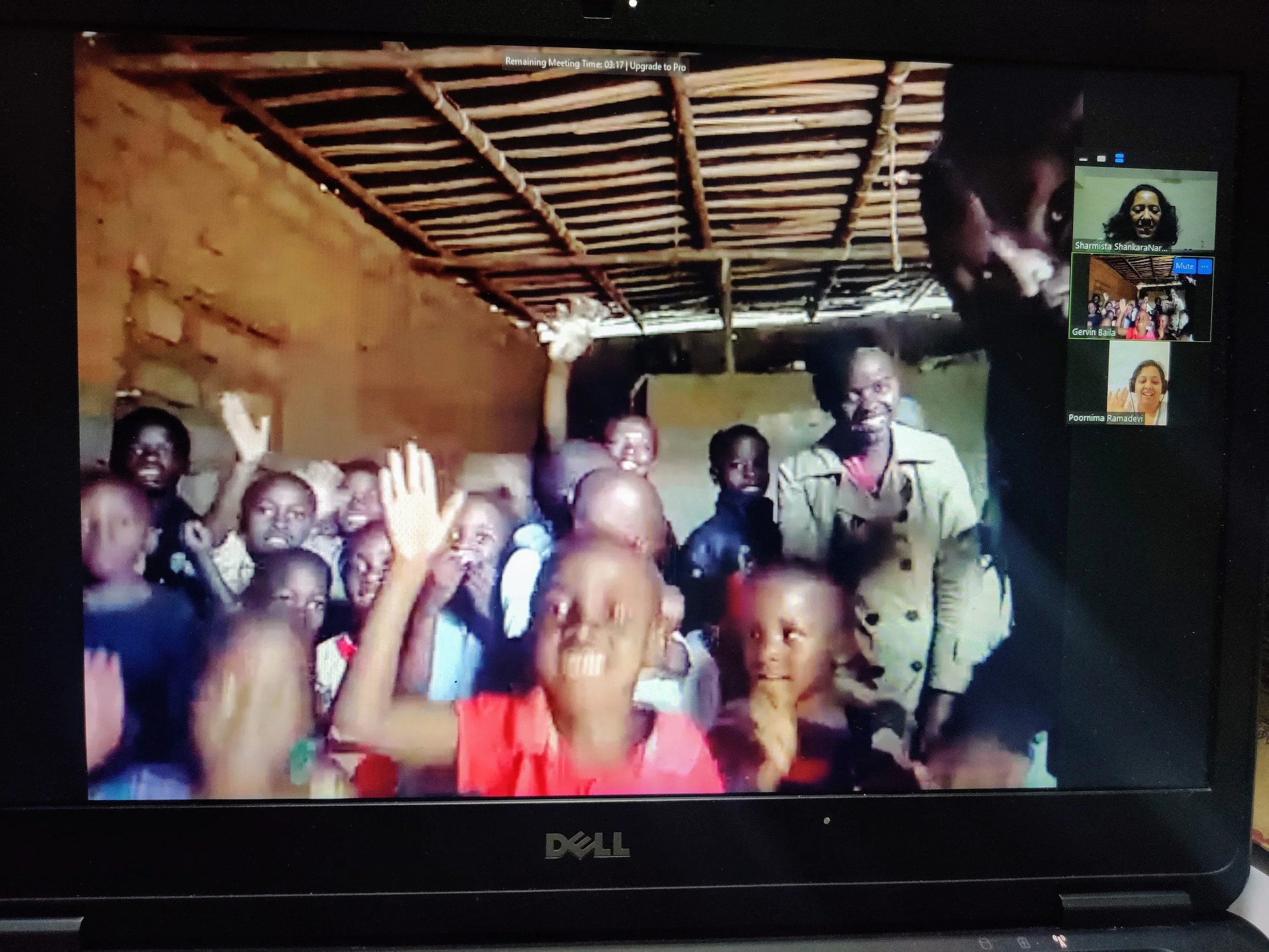 A group of children and an adult inside a rustic room with a thatched roof, some children raising their hands, and others smiling and looking at the camera.