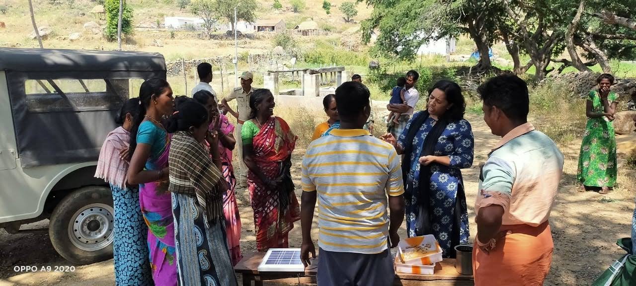 A group of people, including women in colorful traditional Indian sarees, gather outdoors around a woman in a blue patterned dress who is speaking. There are trees and a vehicle in the background, and a table with documents or items on it.