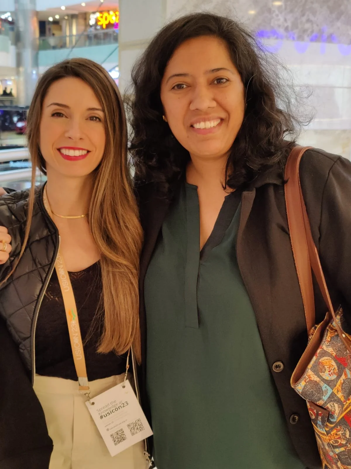 Two women standing close together indoors, smiling at the camera. one has long brown hair and is wearing a black jacket, the other has curly black hair and is wearing a dark green blouse and a black jacket. The woman on the left has a conference badge hanging from her neck.