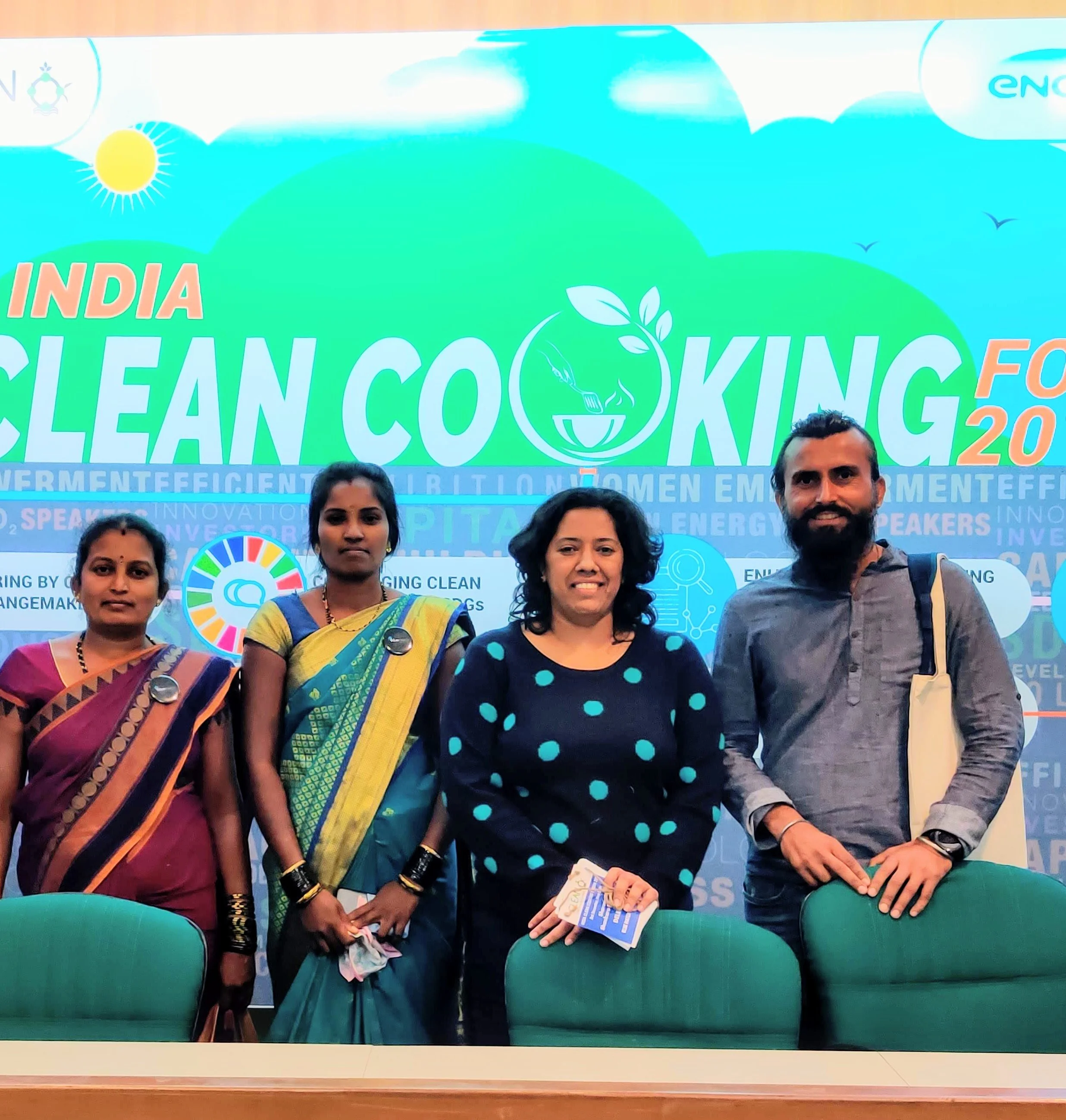 Group of four people standing together at a conference or event, with a banner and audience seats in the background.
