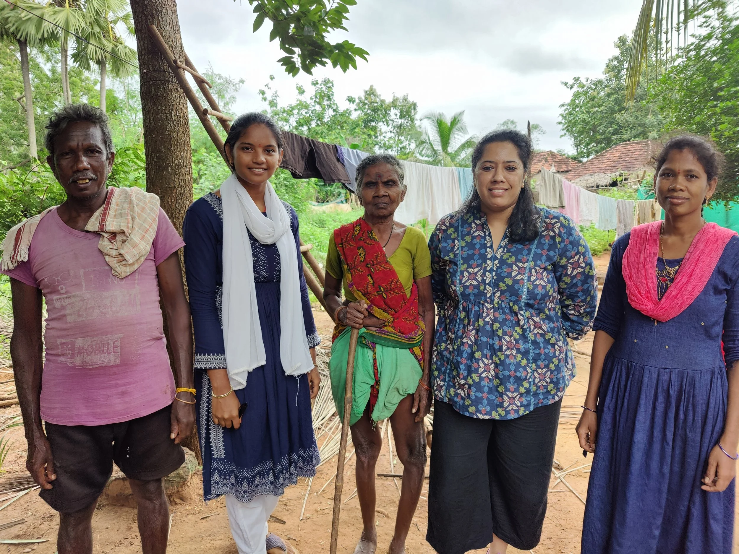 A group of seven women and men stand outdoors in a rural area, surrounded by trees and clothes hanging on a line. They are smiling and appear to be posing for the photo.