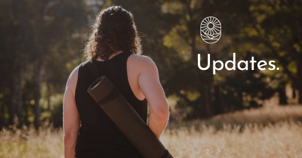 A person seen from behind, a rolled yoga mat slung across their back, walking into dry grass and trees. Text reads "Updates."