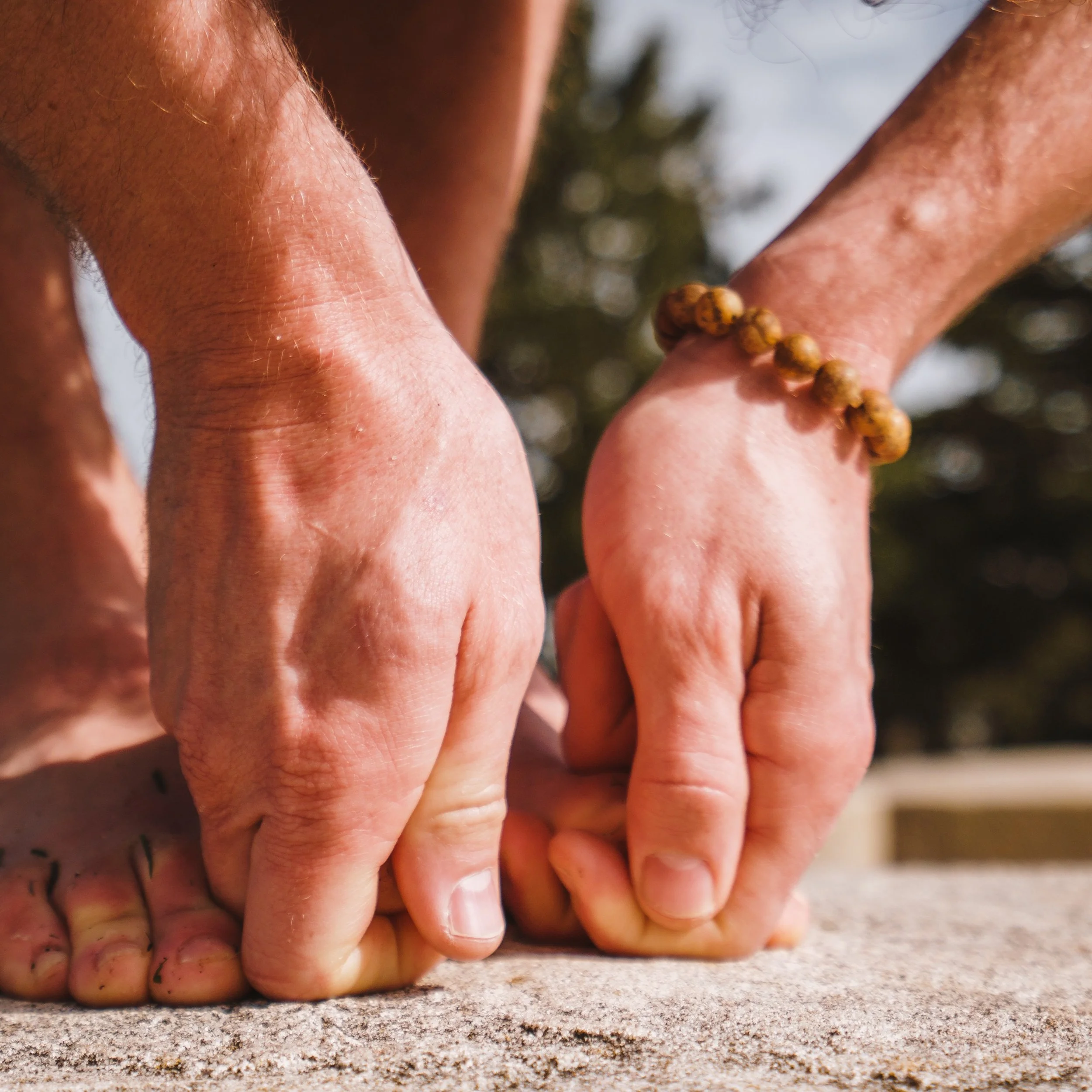 Two hands, one wearing a wooden bead bracelet, press into rough stone beside a bare foot, with trees out of focus behind.