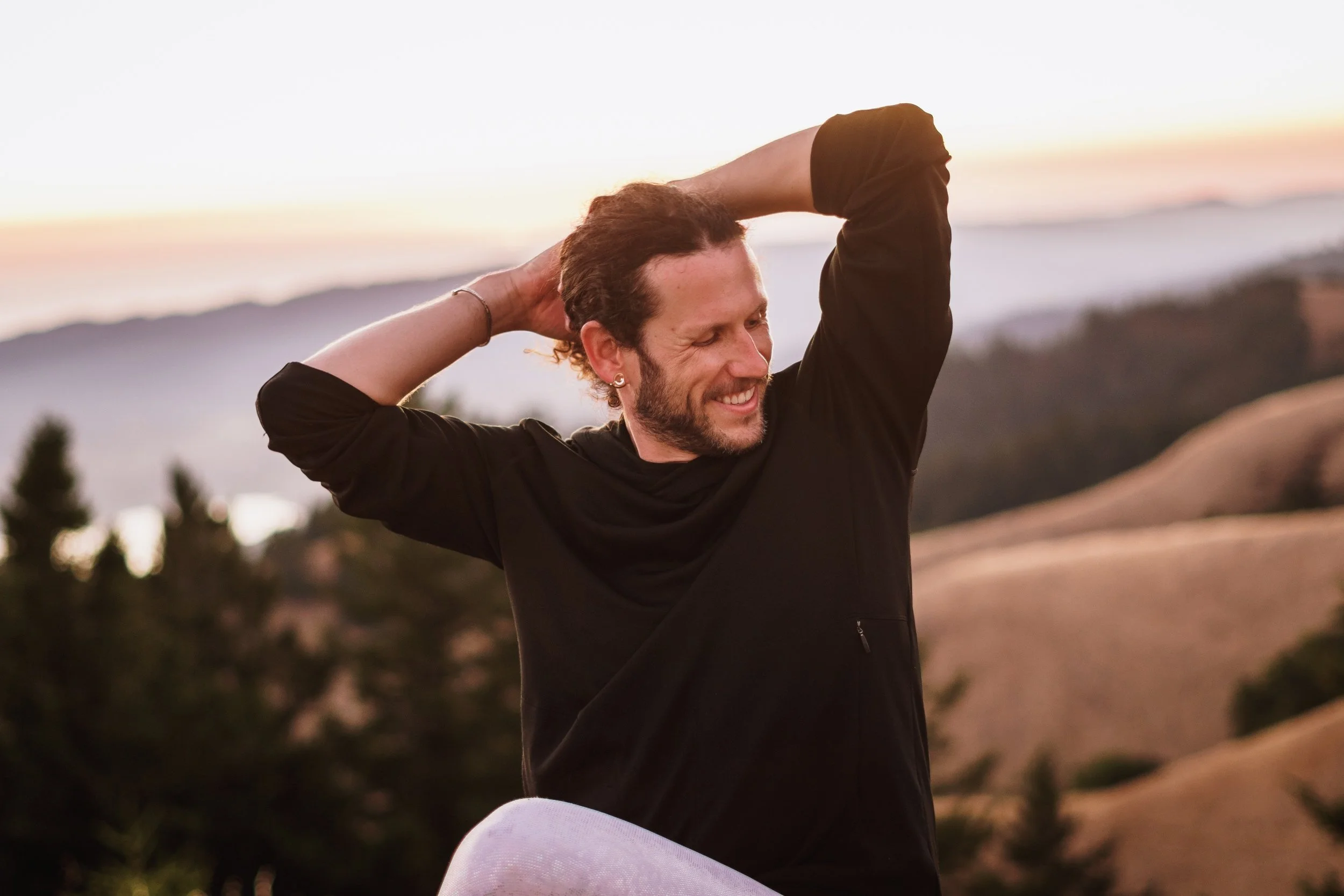 A man smiling outdoors at sunset, with arms raised and hands behind his head, against a background of hills, trees, and a sky with soft pastel colors.