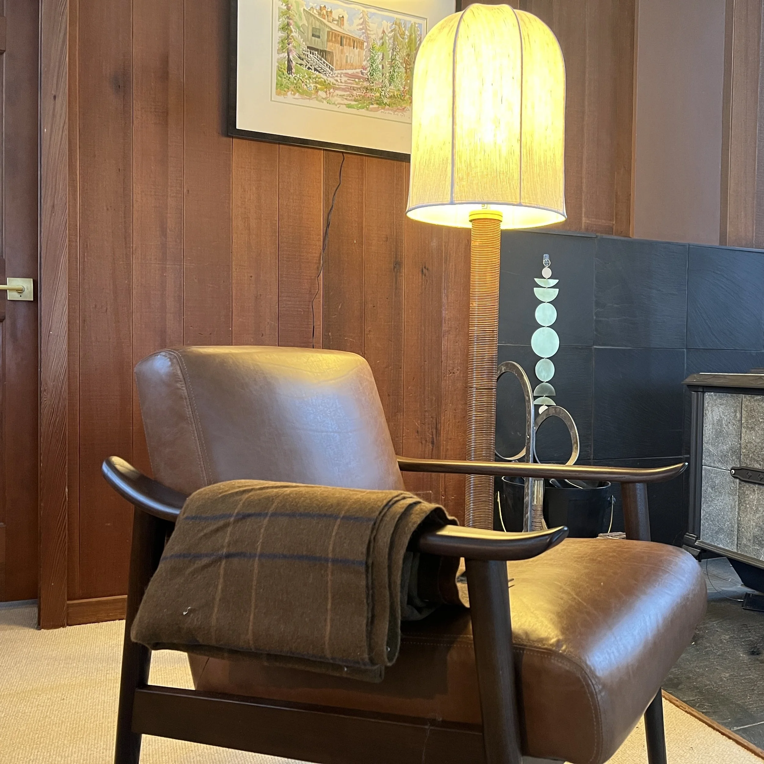 A leather armchair with a folded wool blanket over one arm, beside a lit floor lamp in a wood-panelled room.
