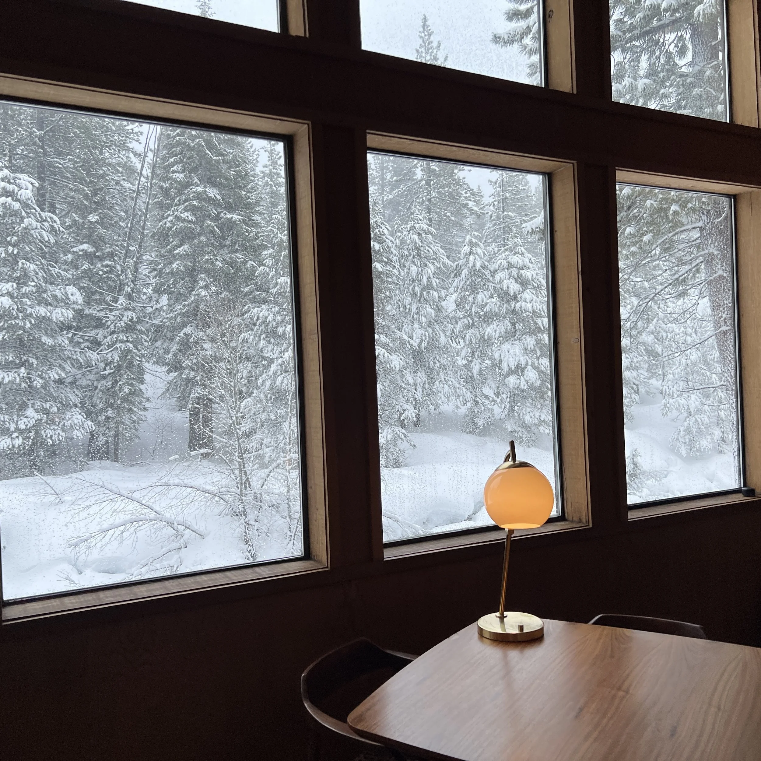 A small lamp glows on a wooden table in front of tall windows looking out on snow-covered pines.