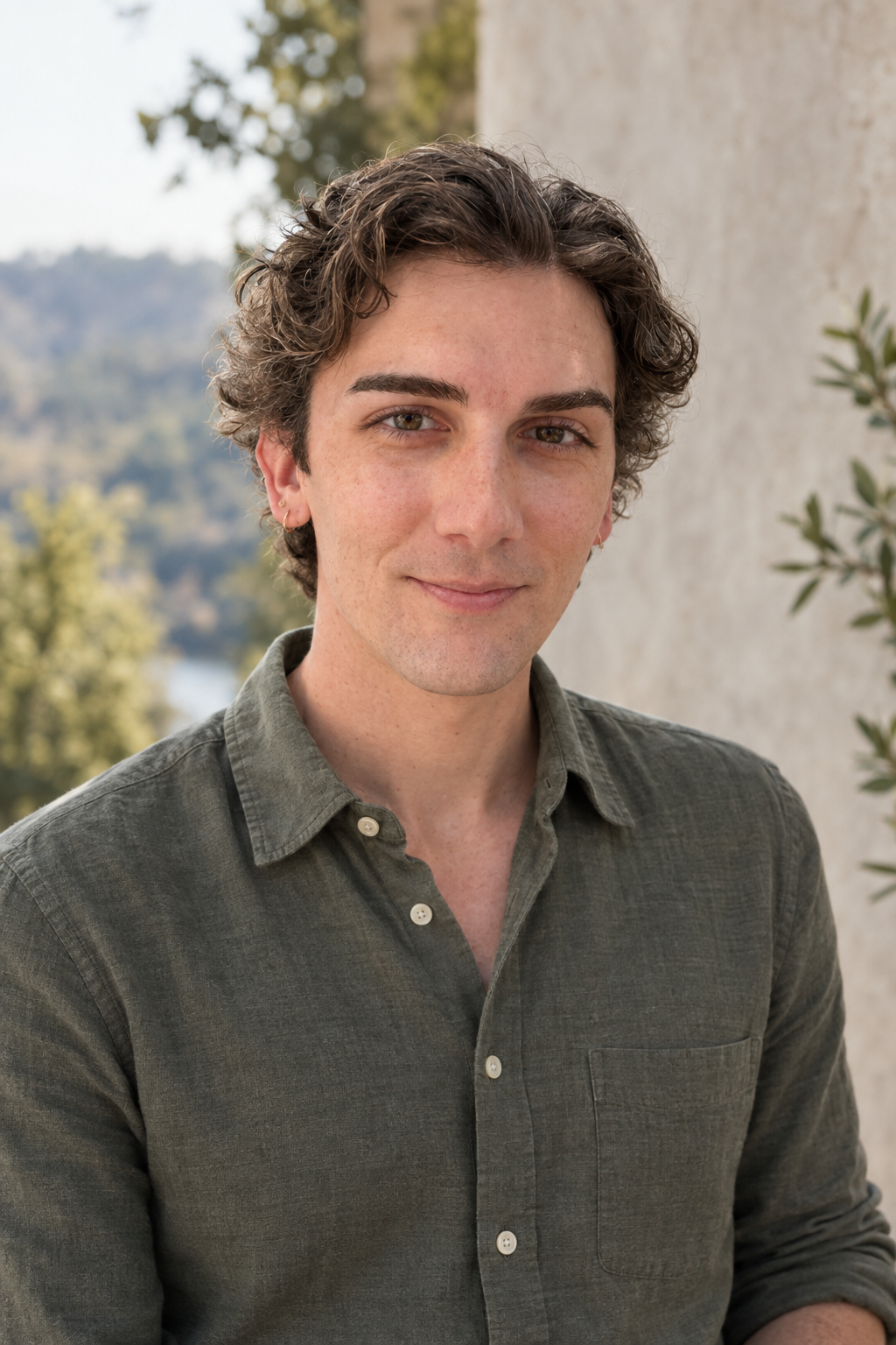 A young man with curly brown hair and earrings, wearing a green button-up shirt, outdoors with trees and a body of water in the background.