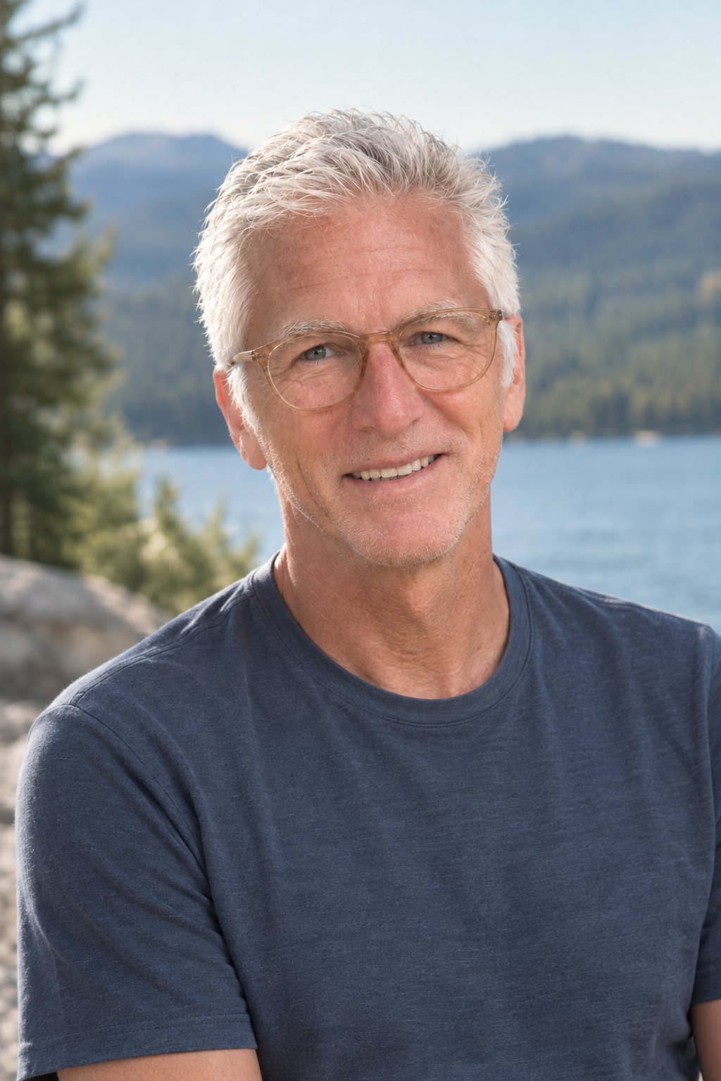 An older man with gray hair and glasses smiling outdoors with a lake, trees, and mountains in the background.