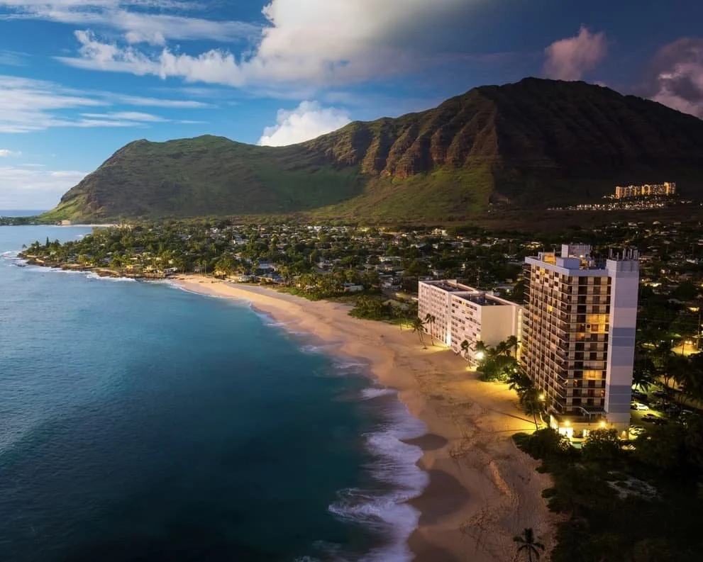 An aerial view of a Hawaiian beach at sunset with a mountain in the background, a hotel along the shoreline, and residential area nearby.
