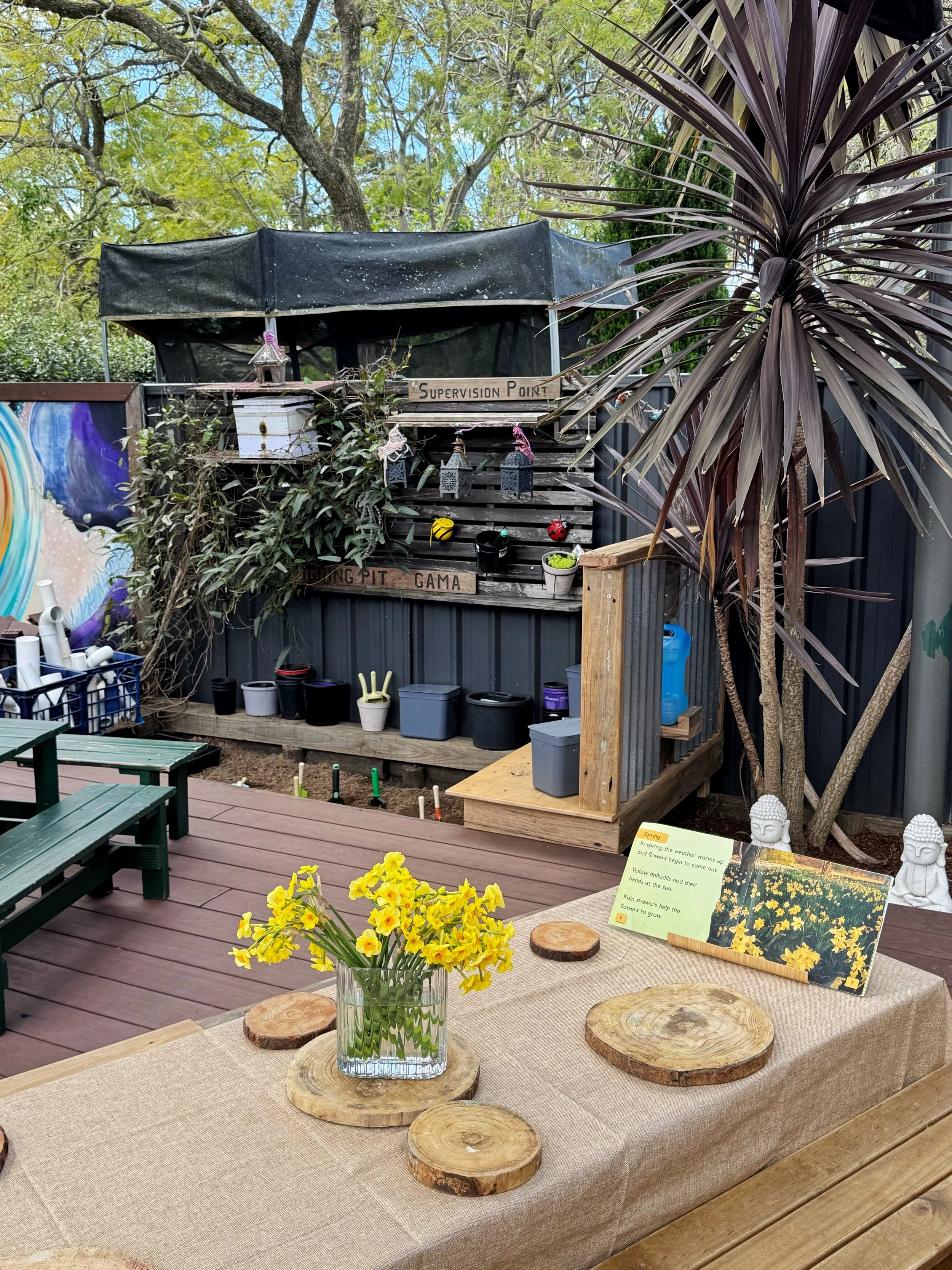 Panania Childcare Centre nature outdoor play - vase of flowers, wooden slices used as coasters. garden area with plants, plant containers and garden tools.