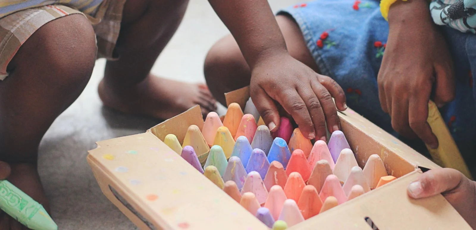 A child's hand reaching into a box of colorful chalks, with other children nearby.