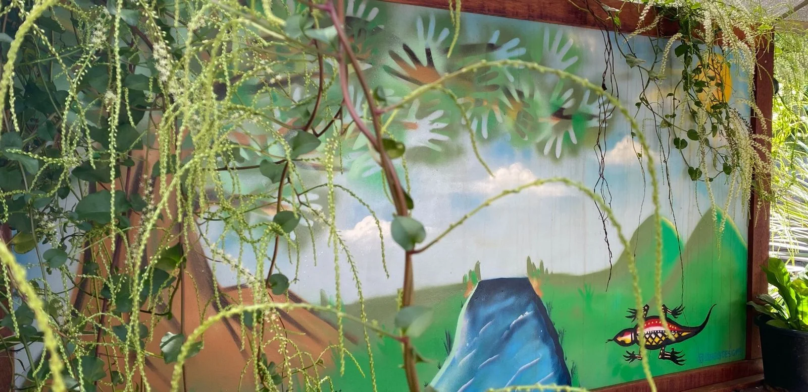 A mural scene with mountains and a colorful lizard in front of a painted landscape, partly obscured by hanging green plants.