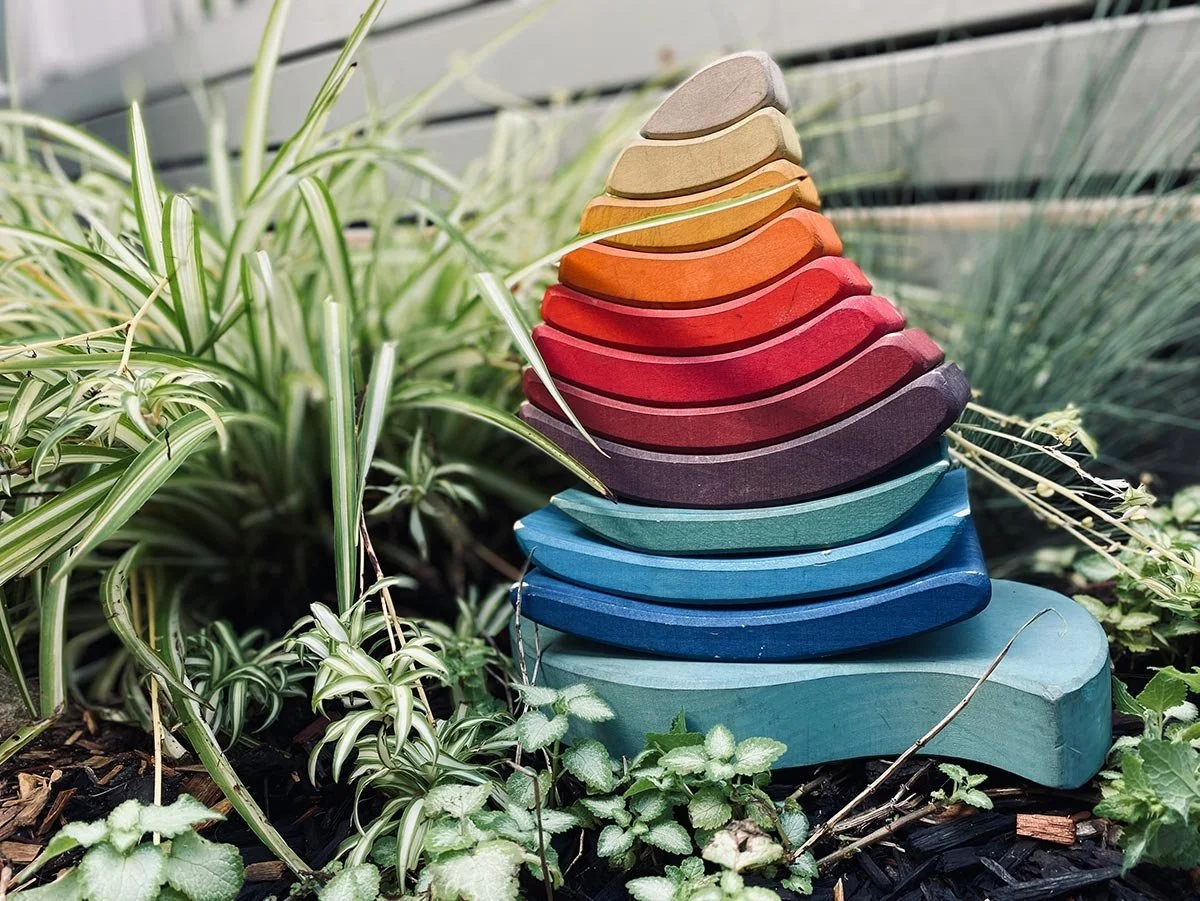 Colorful wooden stacking bird sculpture on garden soil surrounded by green plants and foliage.