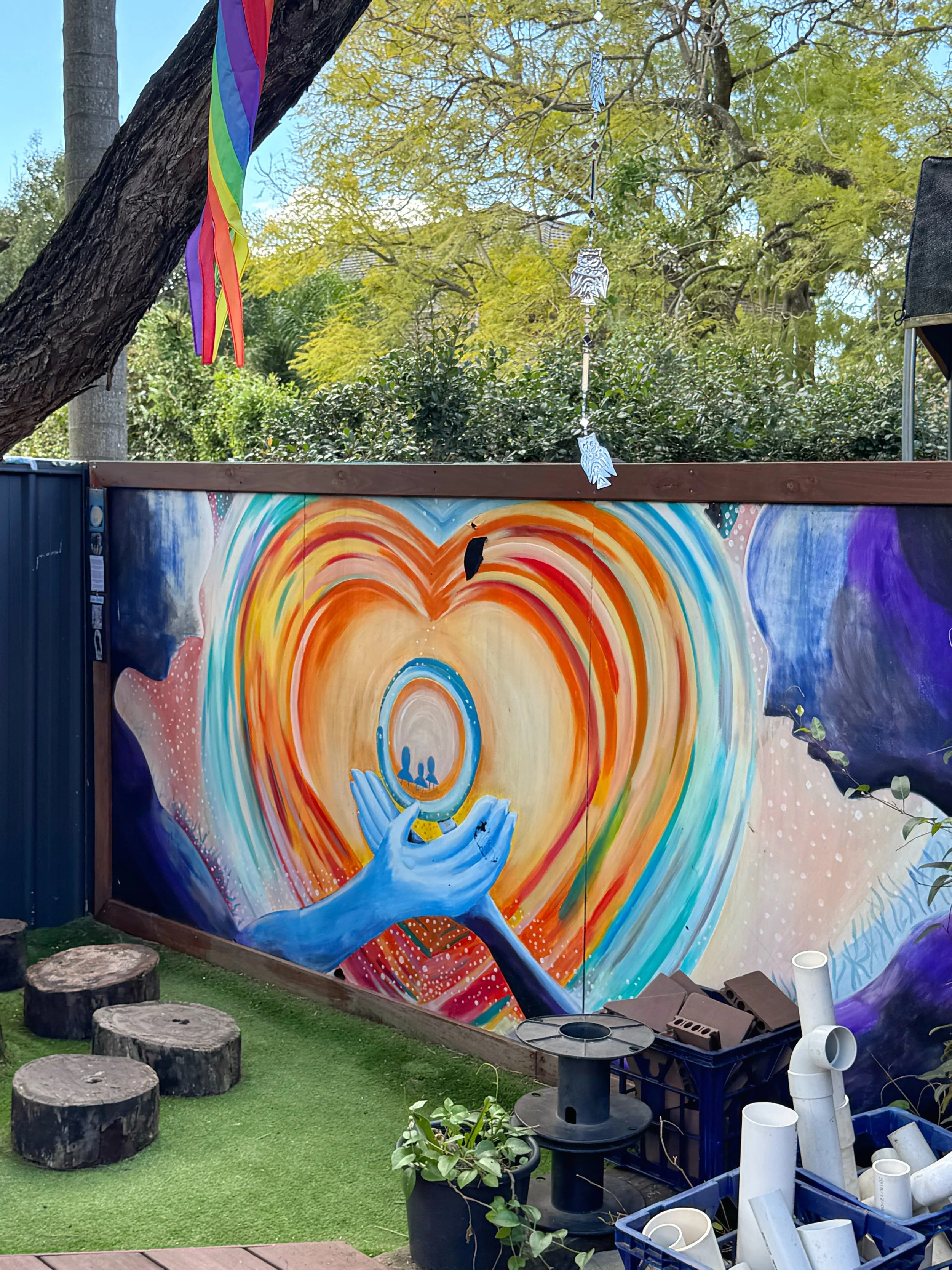 Outdoor learning at Panania Childcare Centre - colourful mural of a heart, backdrop of green trees, rainbow flag hanging from a tree. Various outdoor and creative construction objects such as logs, a blue plastic crate, and PVC pipes.