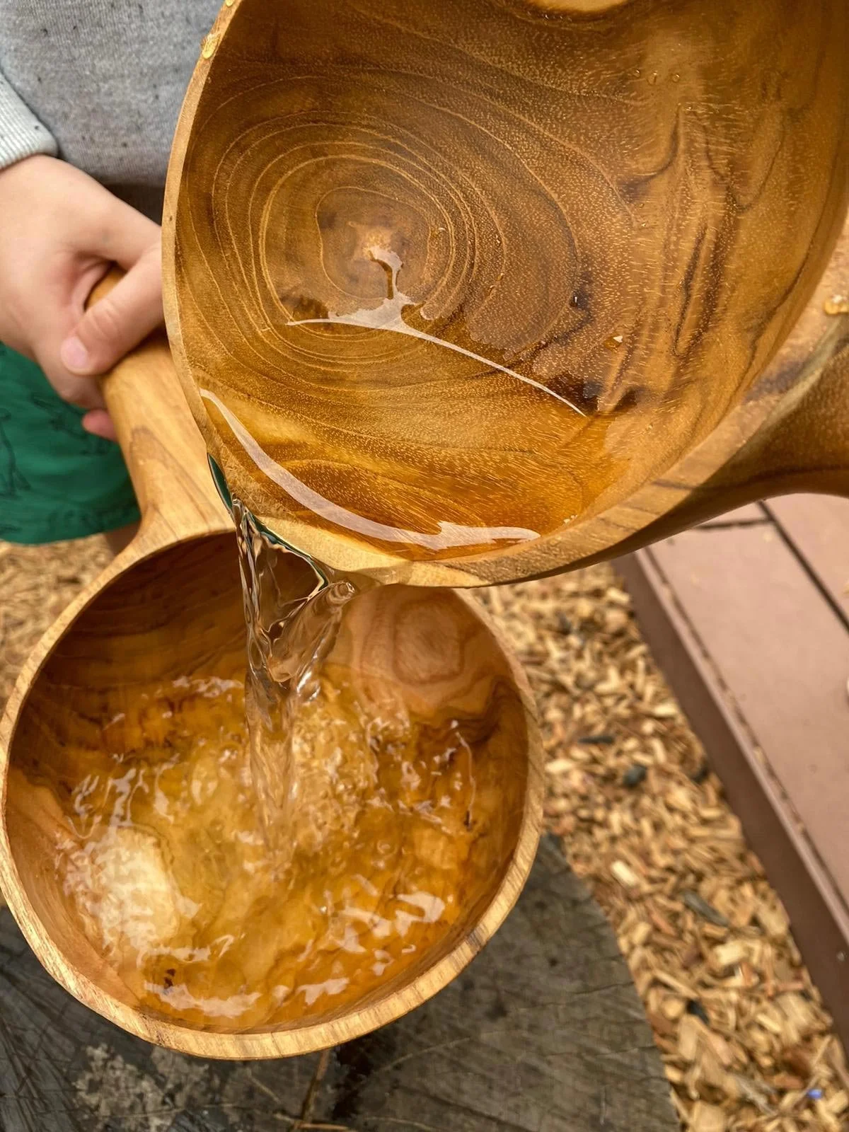 Children pouring water from one wooden bowl into another, with the bowls displaying wood grain patterns, outdoors on a surface with wood mulch.