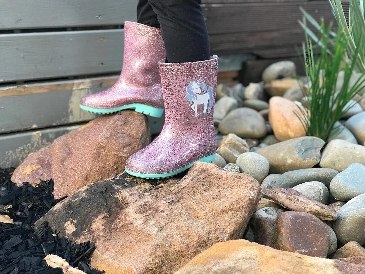 Pink glitter rain boots with a unicorn sticker, placed on rocks near a wooden fence and some plants.