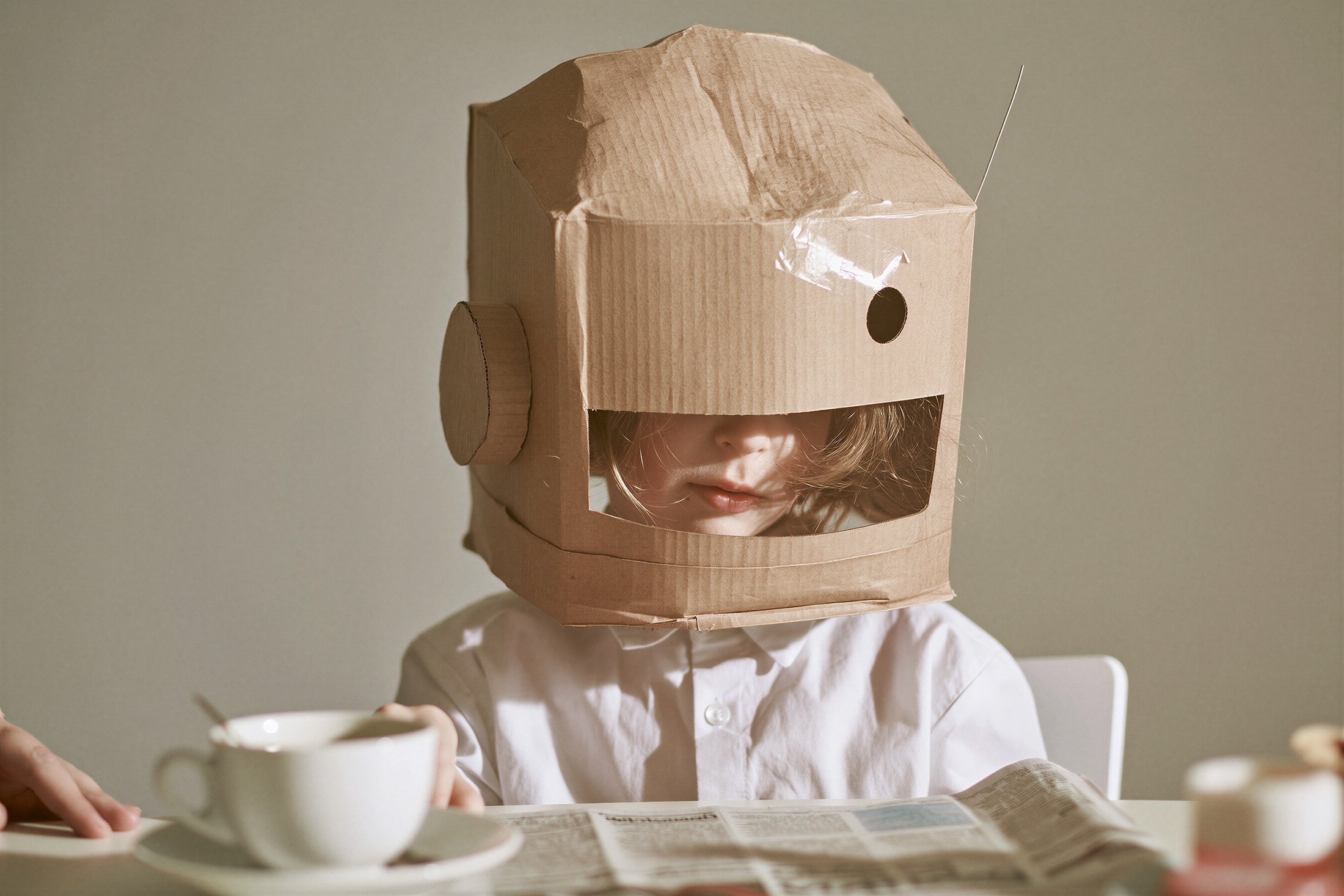Child in a white cotton shirt wearing a handmade cardboard space helmet at a breakfast table with newspaper and teacup — kids advertising. AI-generated image.