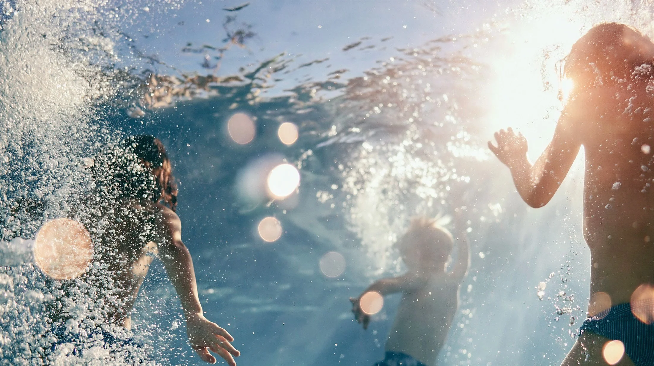 Children underwater against the sun, bubbles and lens flare, swim shorts — kidswear swimwear campaign, backlit, AI-generated image.