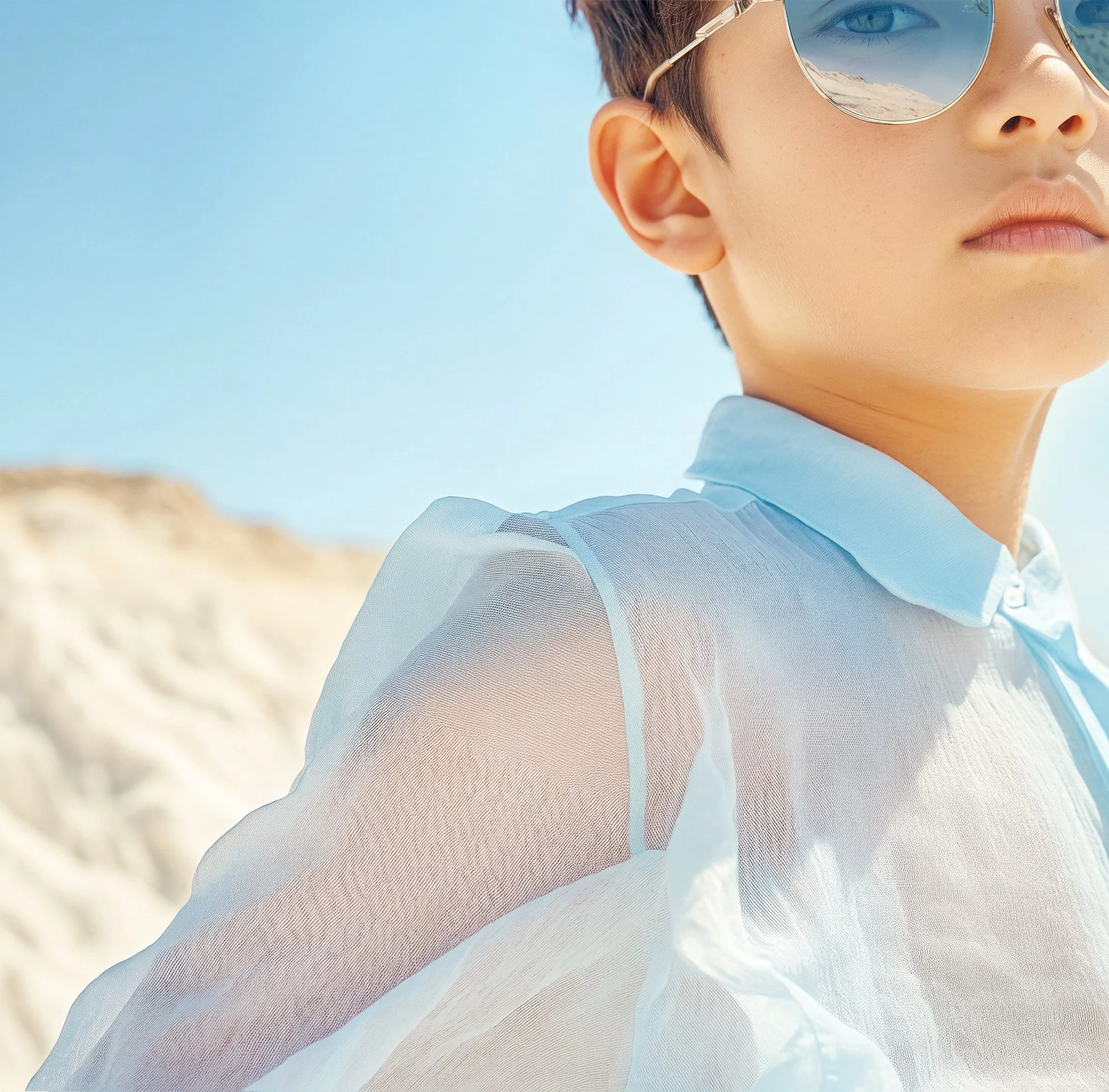 Boy in a sheer blue shirt and mirrored sunglasses against a sunlit dune. AI-generated image.
