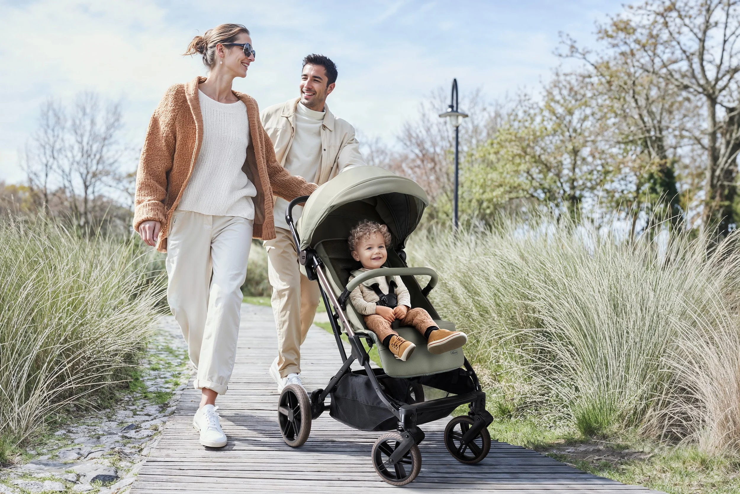 Parents walking a Chicco Dolomiti stroller along a boardwalk through dune grass, toddler smiling in the sage green seat — baby gear campaign for Chicco, family, autumn daylight. AI-generated image.