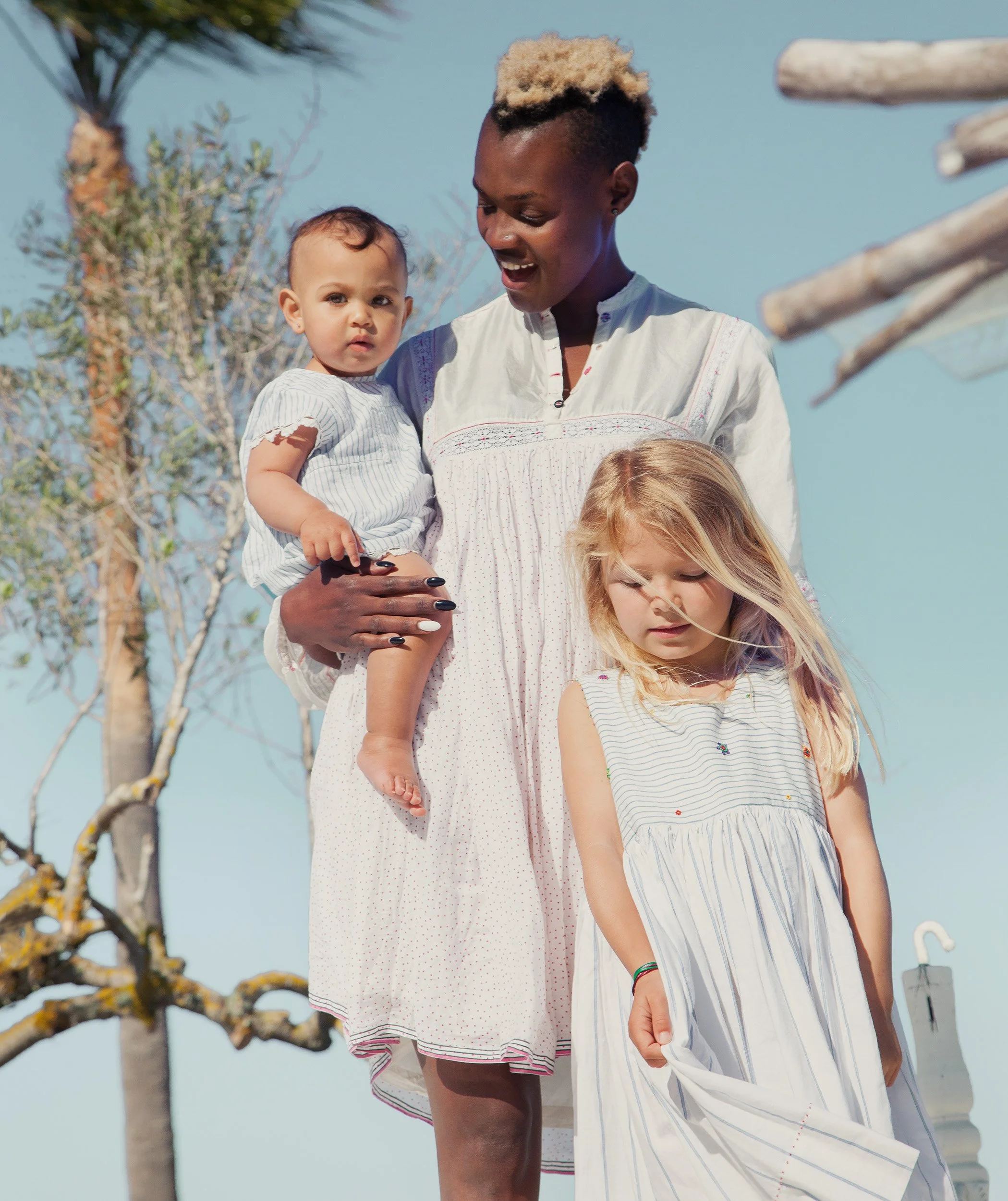 Woman holding a baby beside a small girl, all in coordinated white cotton, against a clear sky — kidswear summer campaign, family, mini-me dressing, diverse casting.