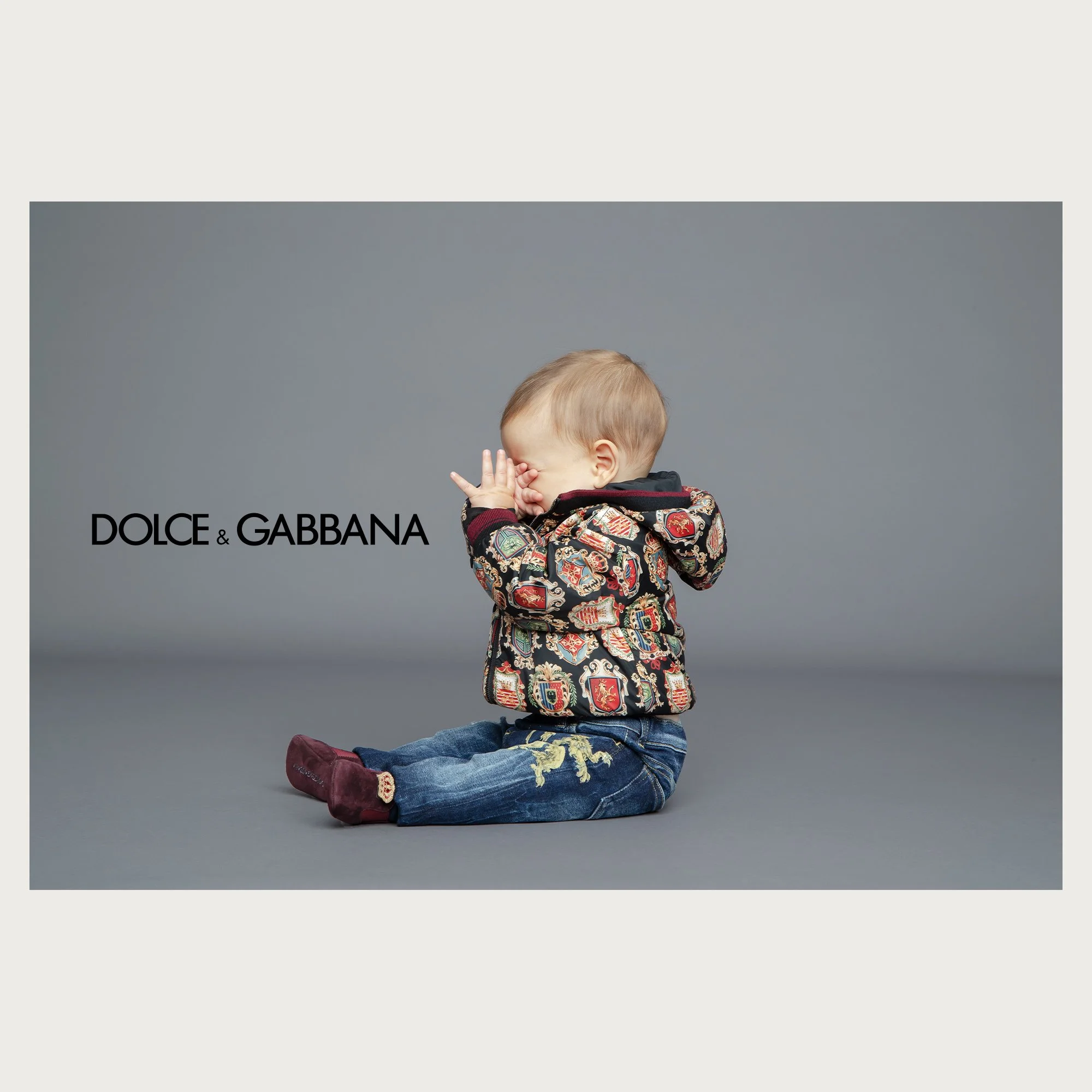 Heraldic print puffer jacket and embroidered jeans, baby covering his face on grey. Dolce & Gabbana newborn campaign.