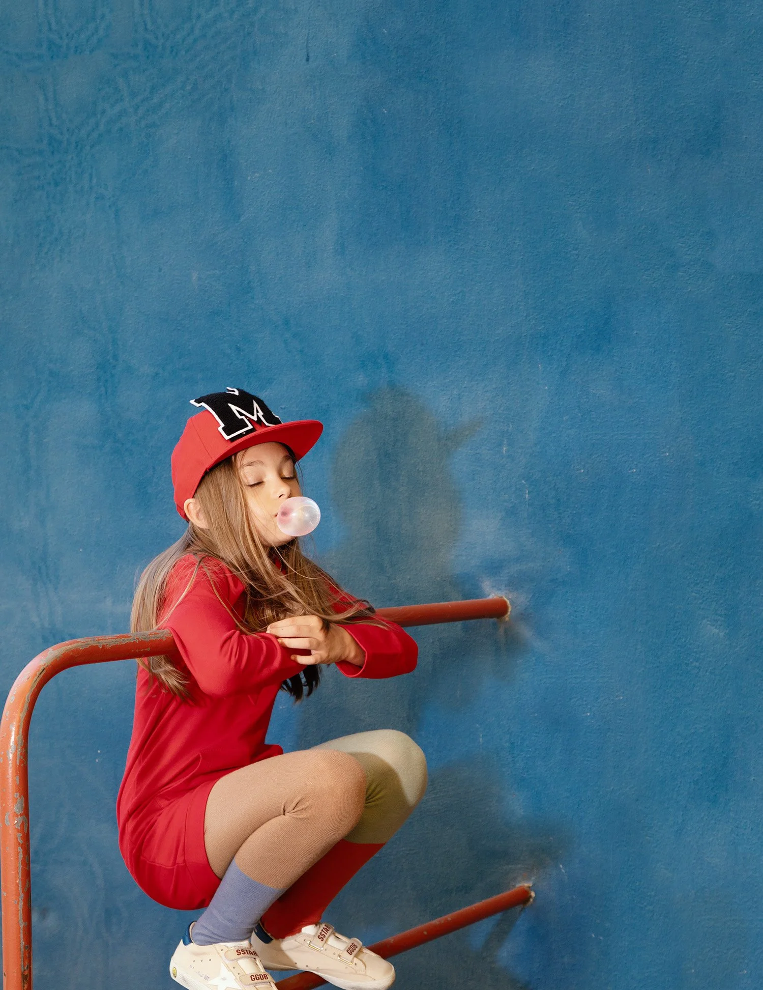 Girl in a red dress and letterman cap blowing a bubblegum bubble, hanging from red railings against a blue wall, eyes closed — kidswear editorial, red on blue.