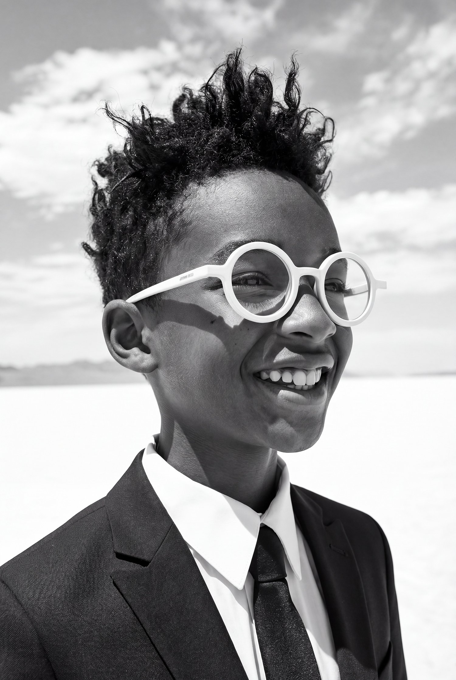 Black and white portrait — boy in a suit and round white glasses, laughing. AI-generated image.