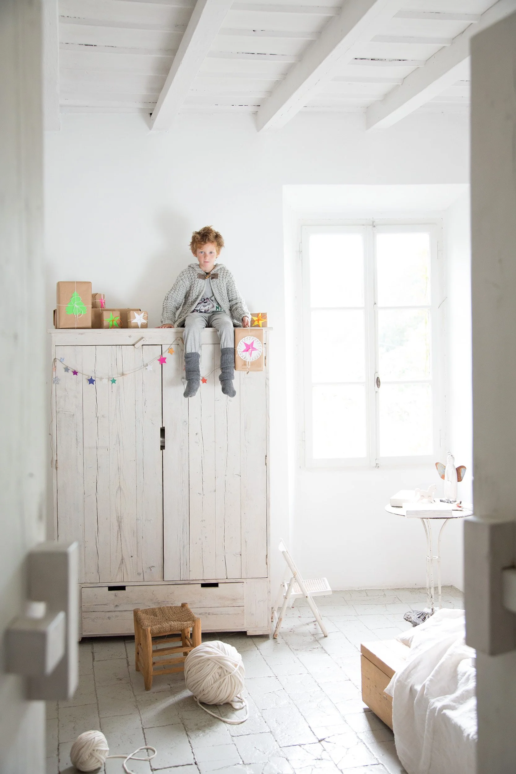 Boy sitting on top of a whitewashed wooden wardrobe in a bright minimal room, white Christmas atmosphere. Style Piccoli editorial.