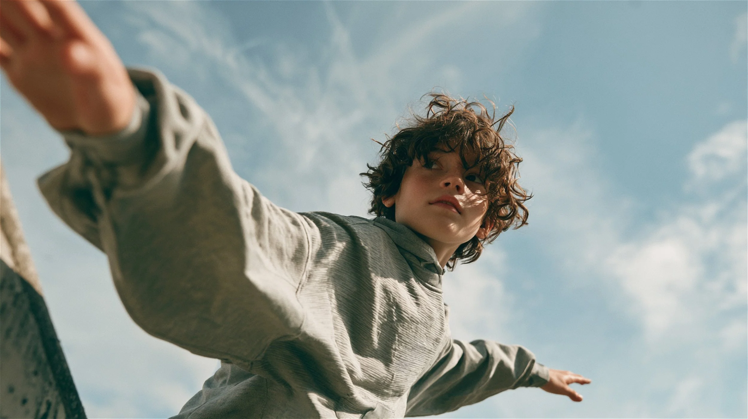 Child with arms out against a blue sky in a sage grey hoodie, seen from below in dappled sunlight — kidswear lifestyle campaign, low angle. AI-generated image.