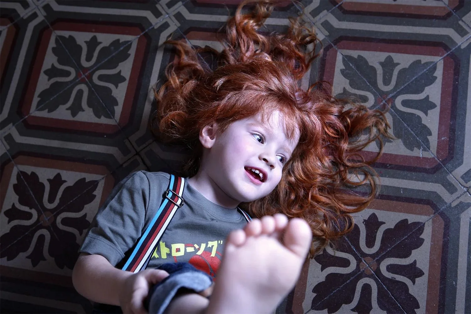Child lying on patterned cement tiles in a grey printed tee and striped suspenders, red curls spread out, bare foot in the foreground — kidswear lifestyle, interiors, high angle.