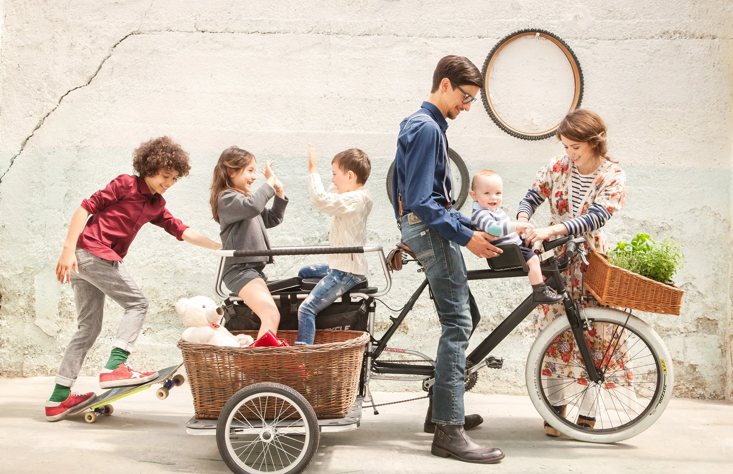 Family of seven with a cargo bike against a concrete wall, five children in shirts, knitwear and denim, skateboard and wicker baskets — kidswear catalogue, group shot.