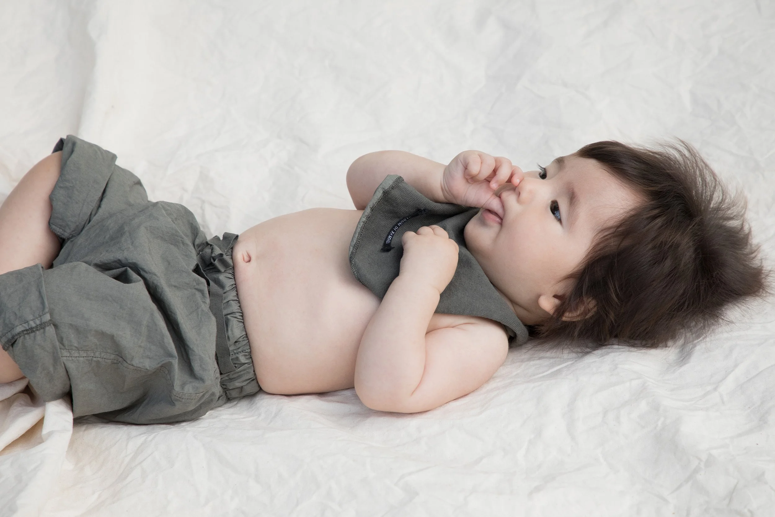 Baby in grey linen shorts and bib on a crumpled white sheet